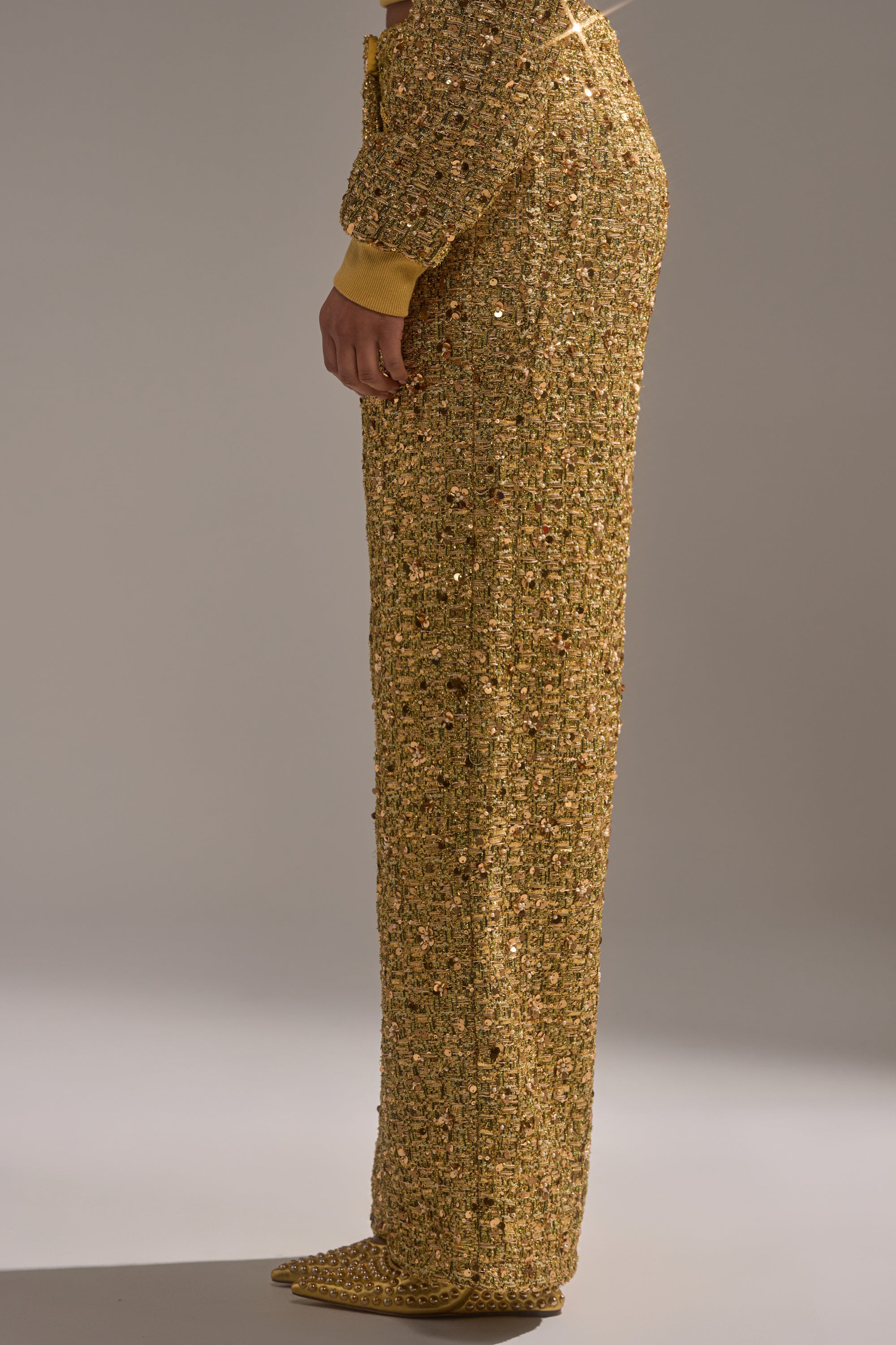 Side view of a person wearing the PRETTY IN GOLD STRAIGHT LEG PANT—gold, textured, sequin-covered trousers with matching pointed shoes—visible from the waist down against a plain, neutral background.
