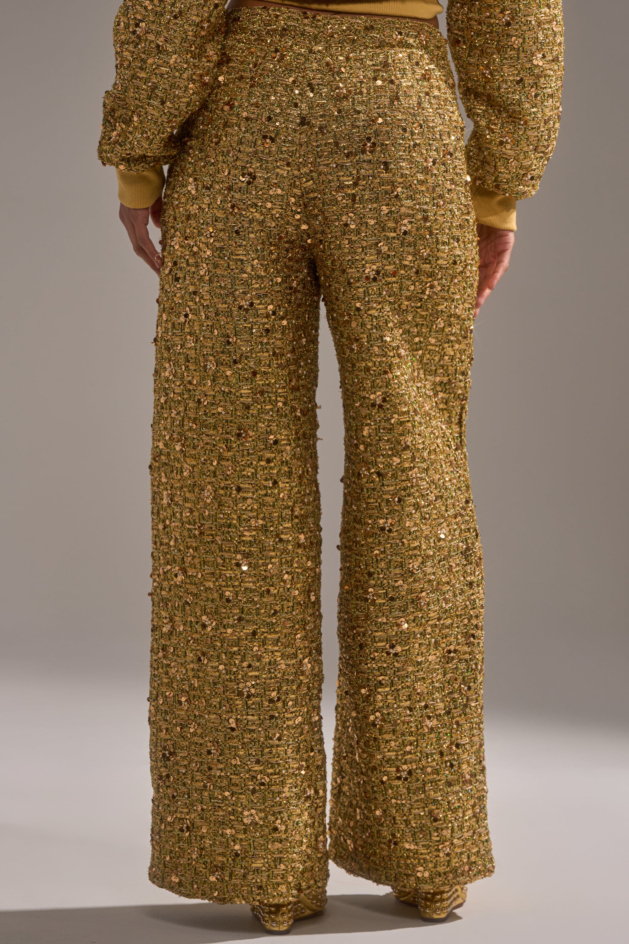 A person faces away, wearing the PRETTY IN GOLD STRAIGHT LEG PANT and a matching textured jacket. The outfit sparkles under the light, showcasing its shimmering golden details.