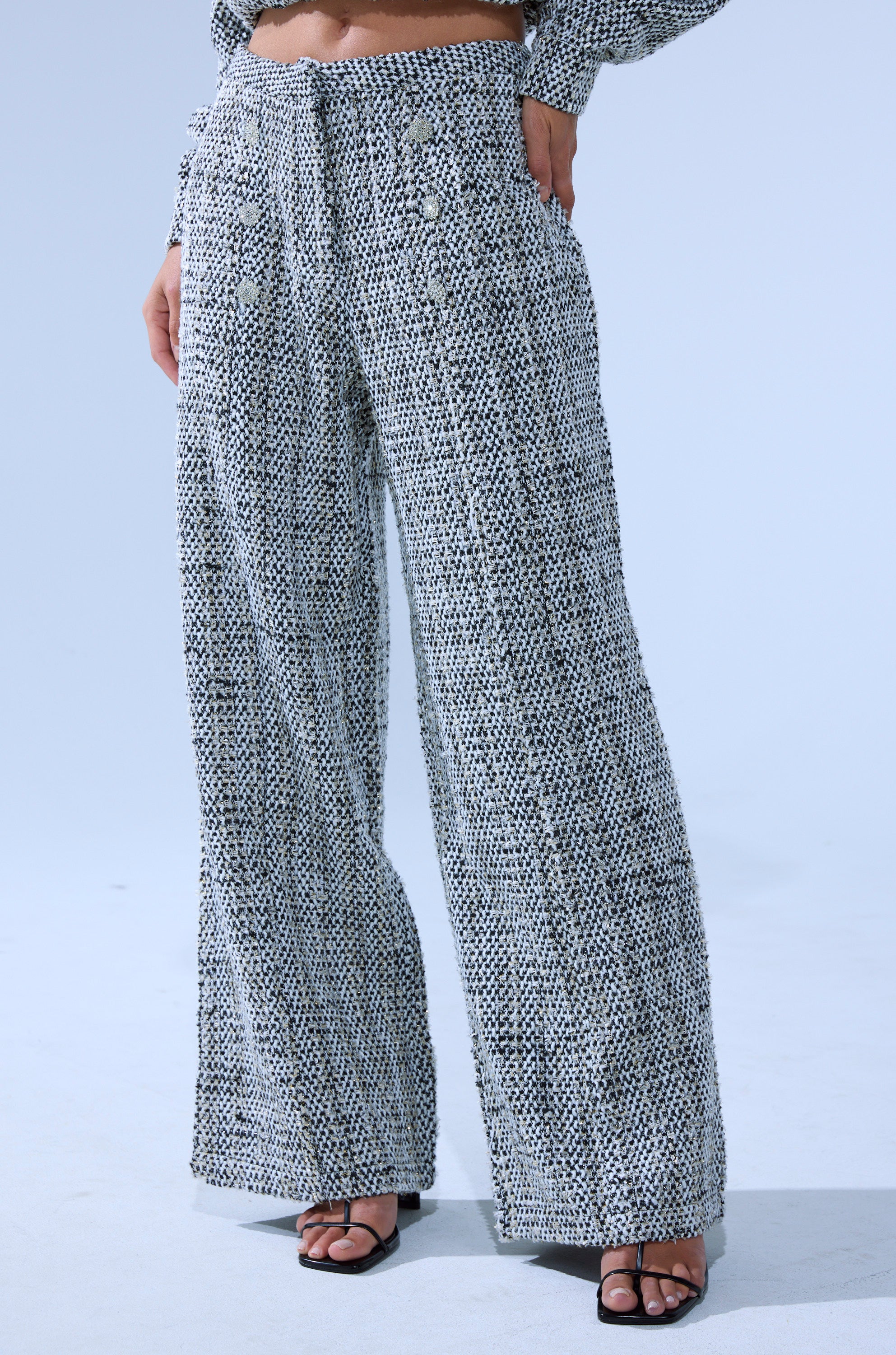 A person models the DRESSY VIBES TWEED PANT, featuring a high-rise wide-leg silhouette with decorative buttons, styled with black open-toe heeled sandals. The background is plain light blue and the upper body is partially visible. 1