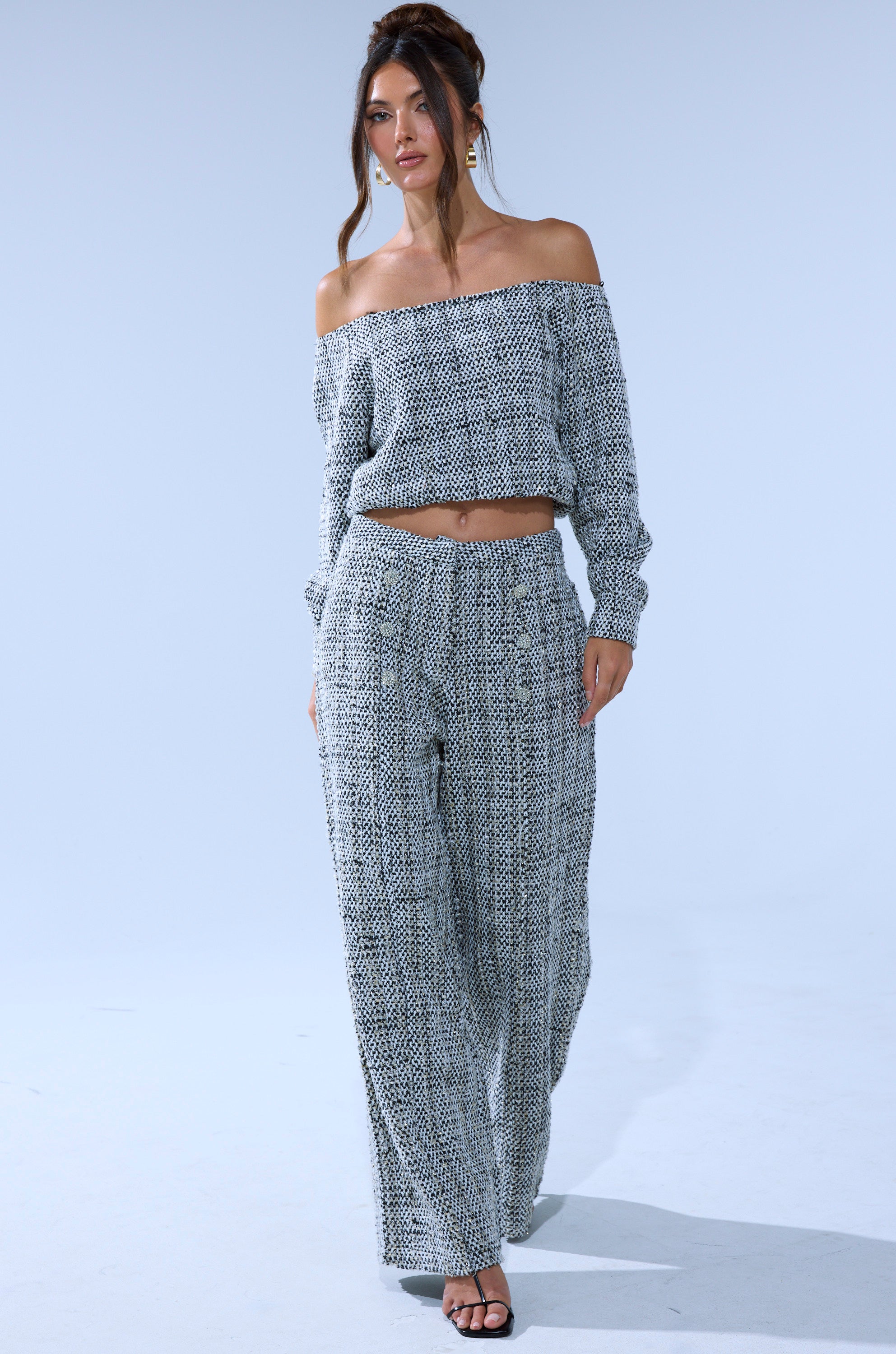 A woman walks forward in black heeled sandals wearing the DRESSY VIBES TWEED PANT, a high-rise, wide-leg silhouette paired with an off-the-shoulder, long-sleeve patterned top against a plain light background. 5