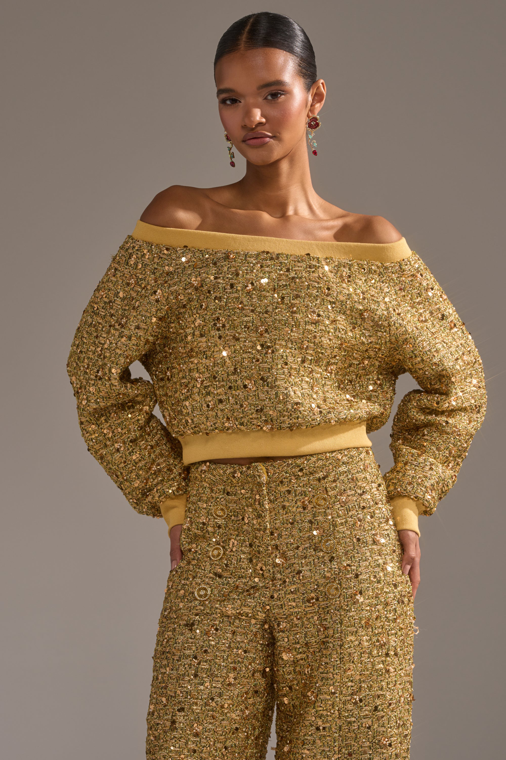 A woman confidently poses with her hands on her hips, wearing the PRETTY IN GOLD OFF THE SHOULDER SWEATSHIRT and matching high-waisted pants against a neutral background.