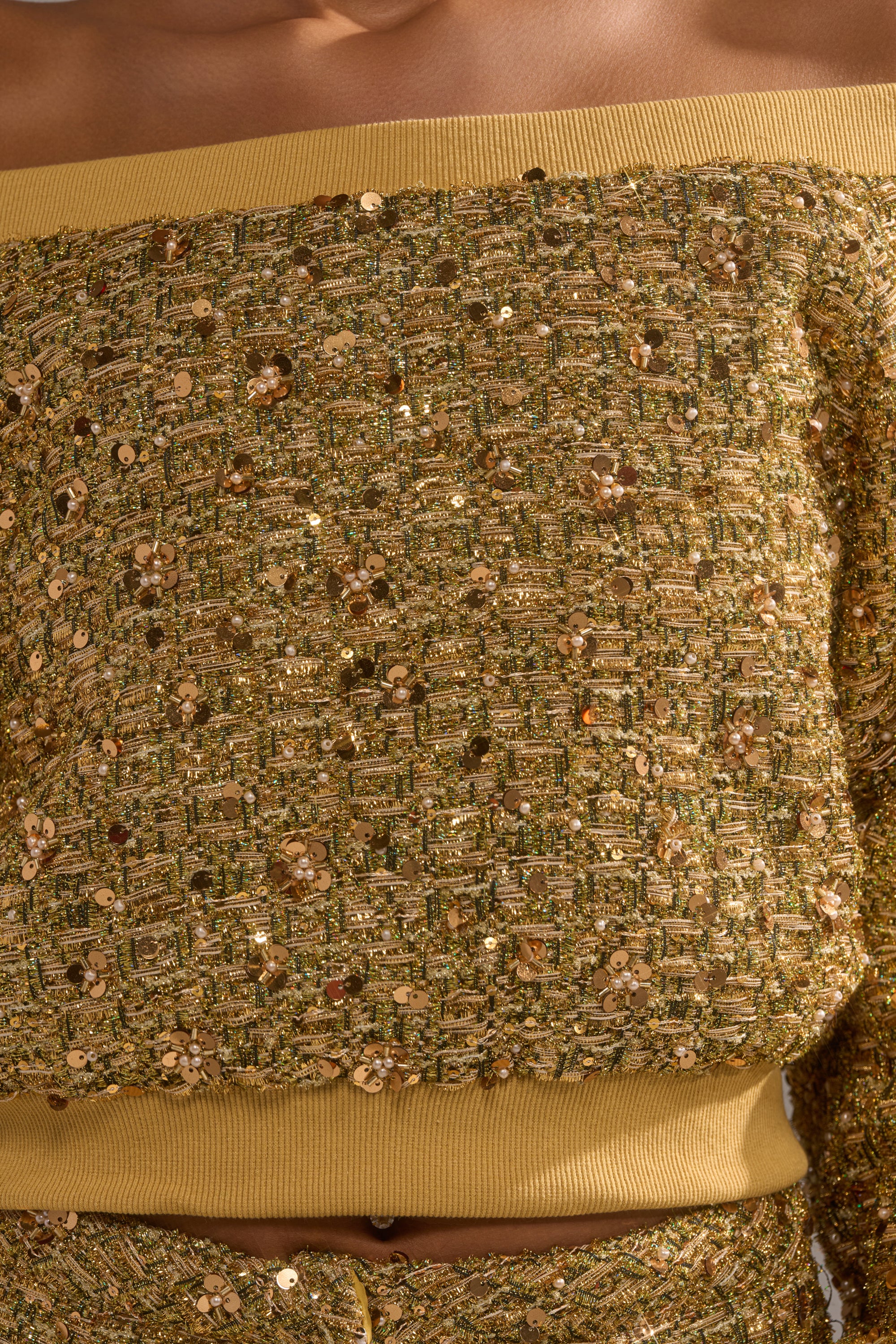 Close-up of a person wearing the PRETTY IN GOLD OFF THE SHOULDER SWEATSHIRT, made from textured gold fabric with sequins and metallic threads, featuring ribbed golden bands at the neckline and hem.