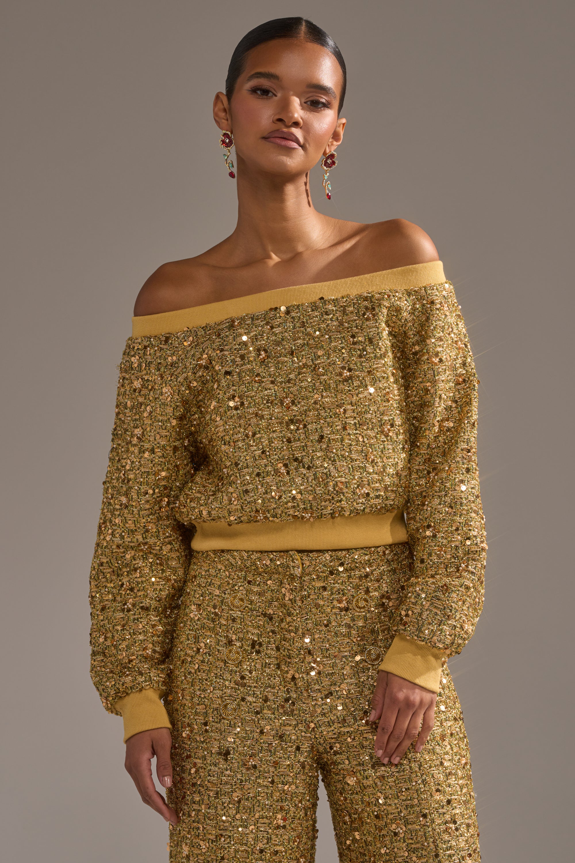 A woman models the PRETTY IN GOLD OFF THE SHOULDER SWEATSHIRT with matching pants, her hair tied back and styled with colorful drop earrings, standing against a plain background.
