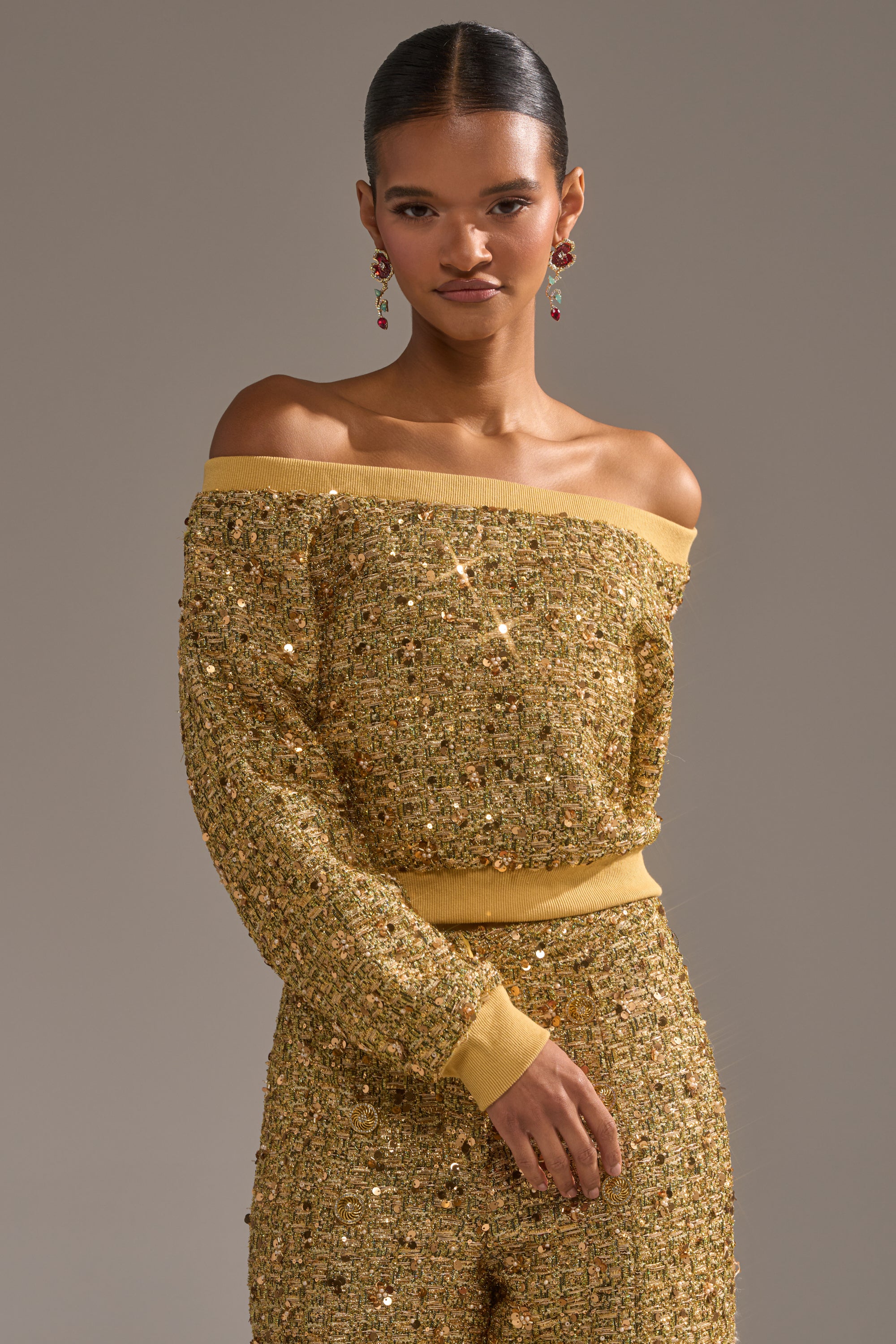 A person wears the PRETTY IN GOLD OFF THE SHOULDER SWEATSHIRT with matching pants, their hair pulled back and accessorized with dangling earrings, standing against a plain background.