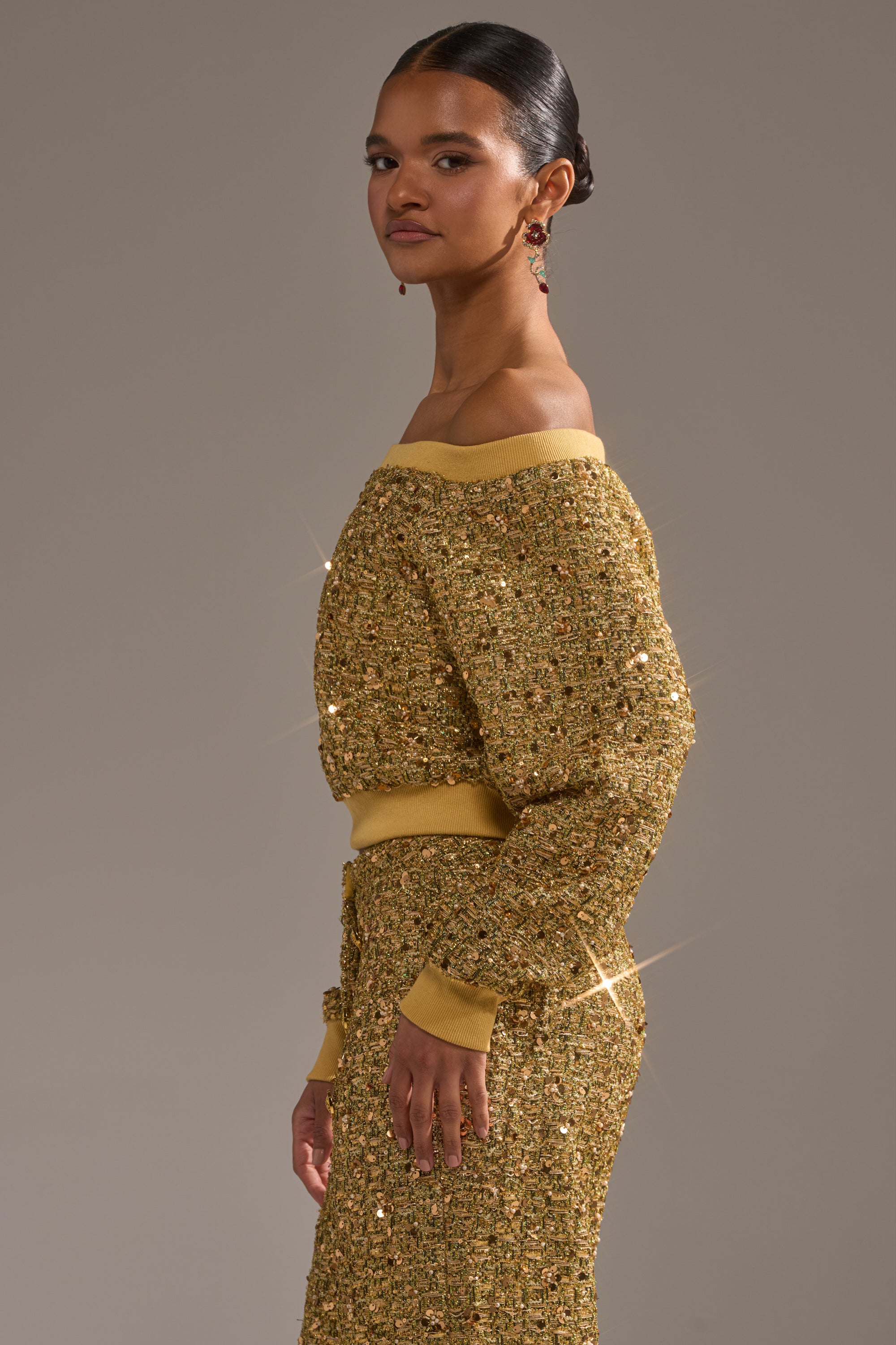A woman stands sideways in the PRETTY IN GOLD OFF THE SHOULDER SWEATSHIRT, a sparkling gold sequin top paired with a matching skirt and dangling earrings against a plain, neutral background.