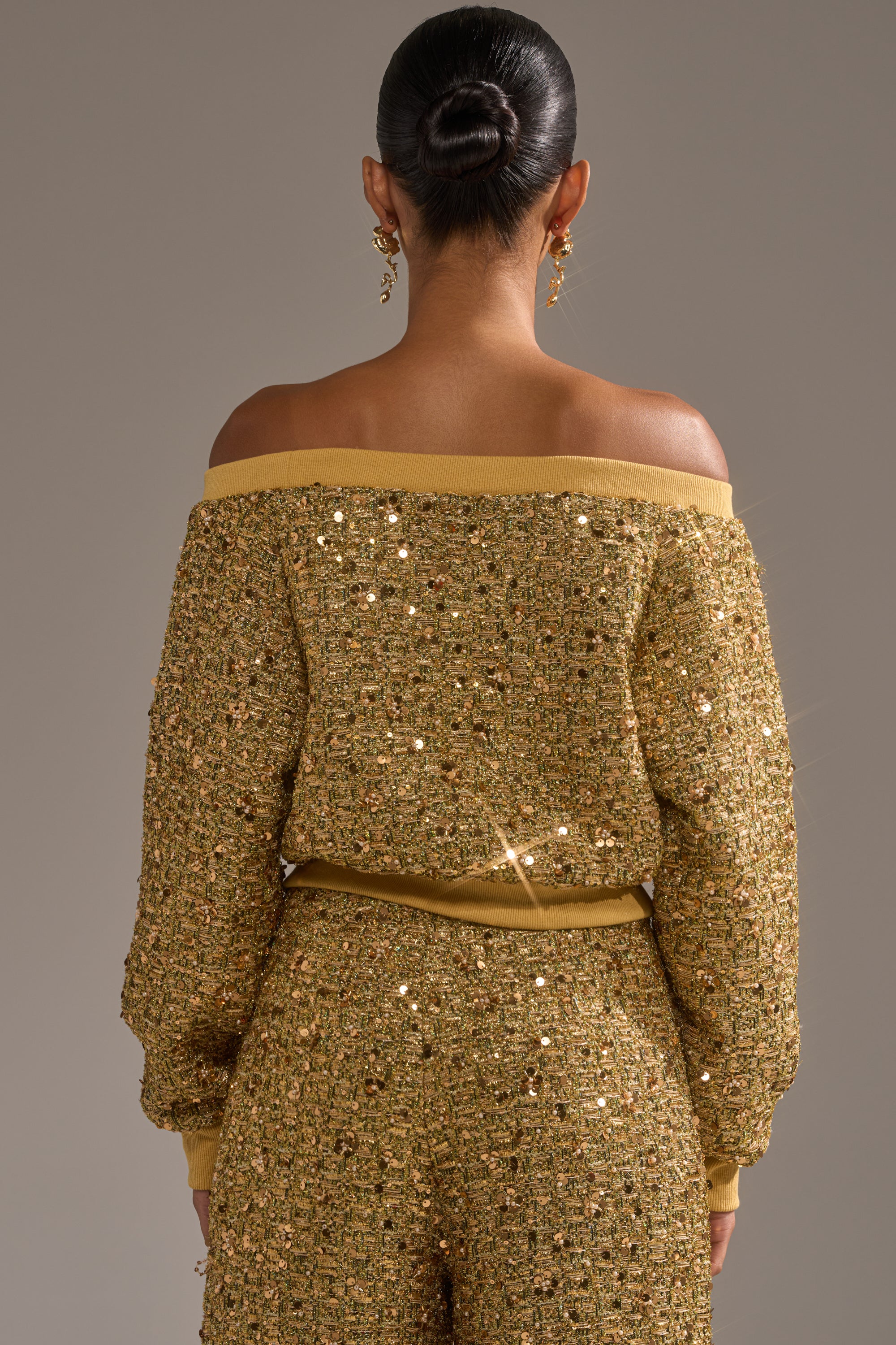 A person with their hair in a sleek bun is shown from behind, wearing the PRETTY IN GOLD OFF THE SHOULDER SWEATSHIRT and matching sequin-embellished pants that shimmer under the light.
