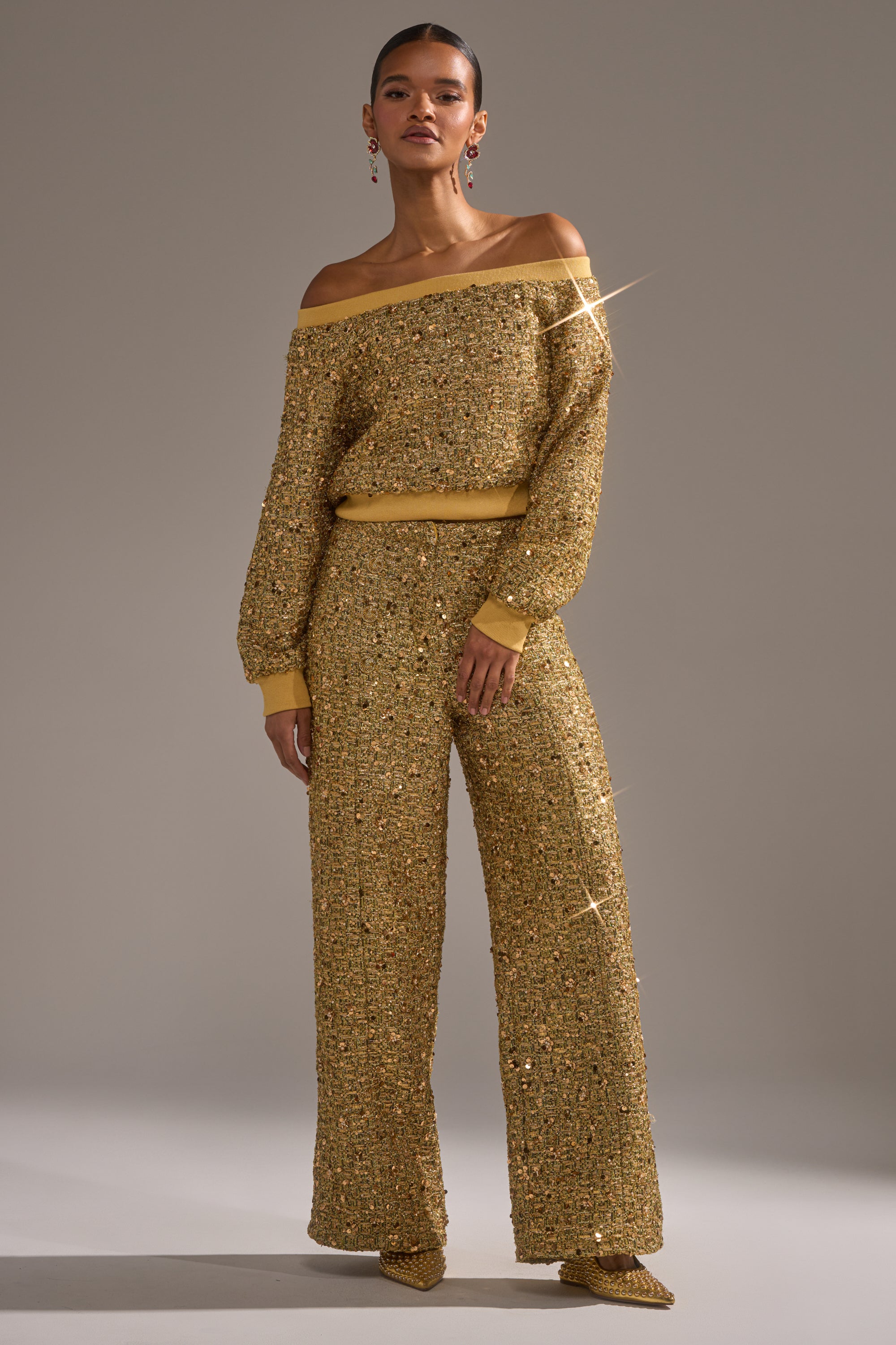 A woman stands confidently against a plain background, wearing the PRETTY IN GOLD OFF THE SHOULDER SWEATSHIRT paired with matching wide-leg pants and pointed shoes.