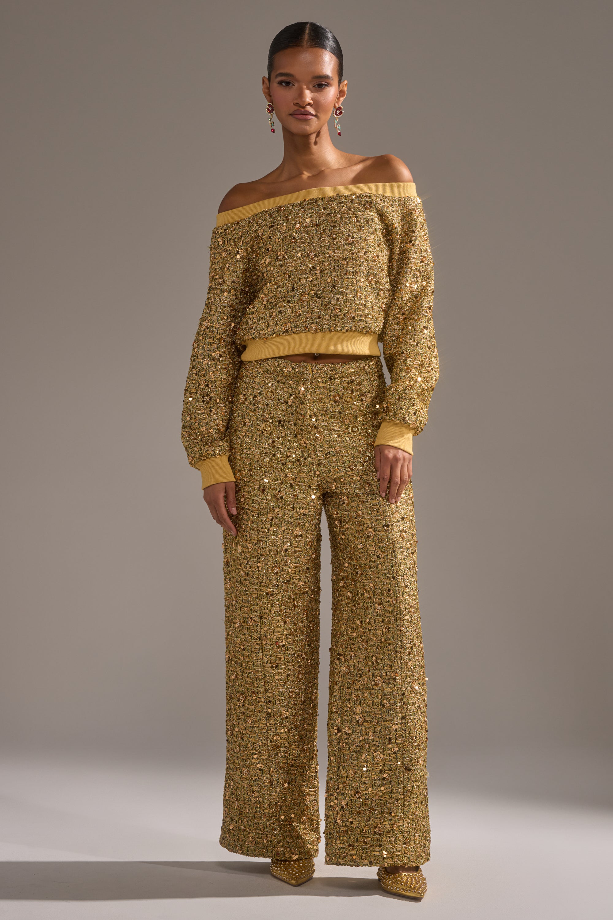 A woman wears the PRETTY IN GOLD OFF THE SHOULDER SWEATSHIRT and matching embellished wide-leg pants with gold shoes and statement earrings, standing against a plain gray background.