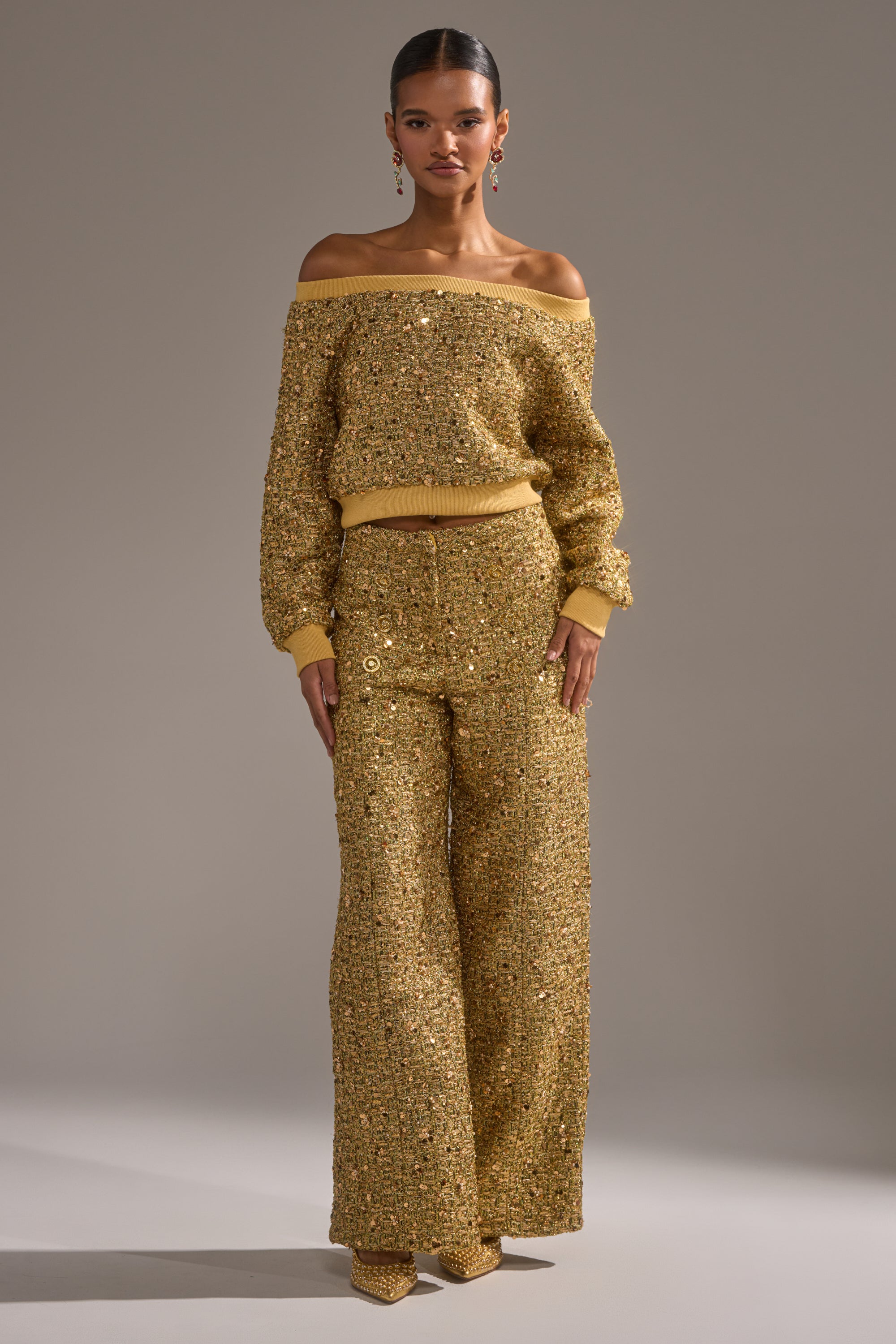 A woman faces forward in the PRETTY IN GOLD OFF THE SHOULDER SWEATSHIRT, paired with matching wide-leg pants and pointed shoes. She wears drop earrings, has slicked-back hair, and stands against a plain neutral background.