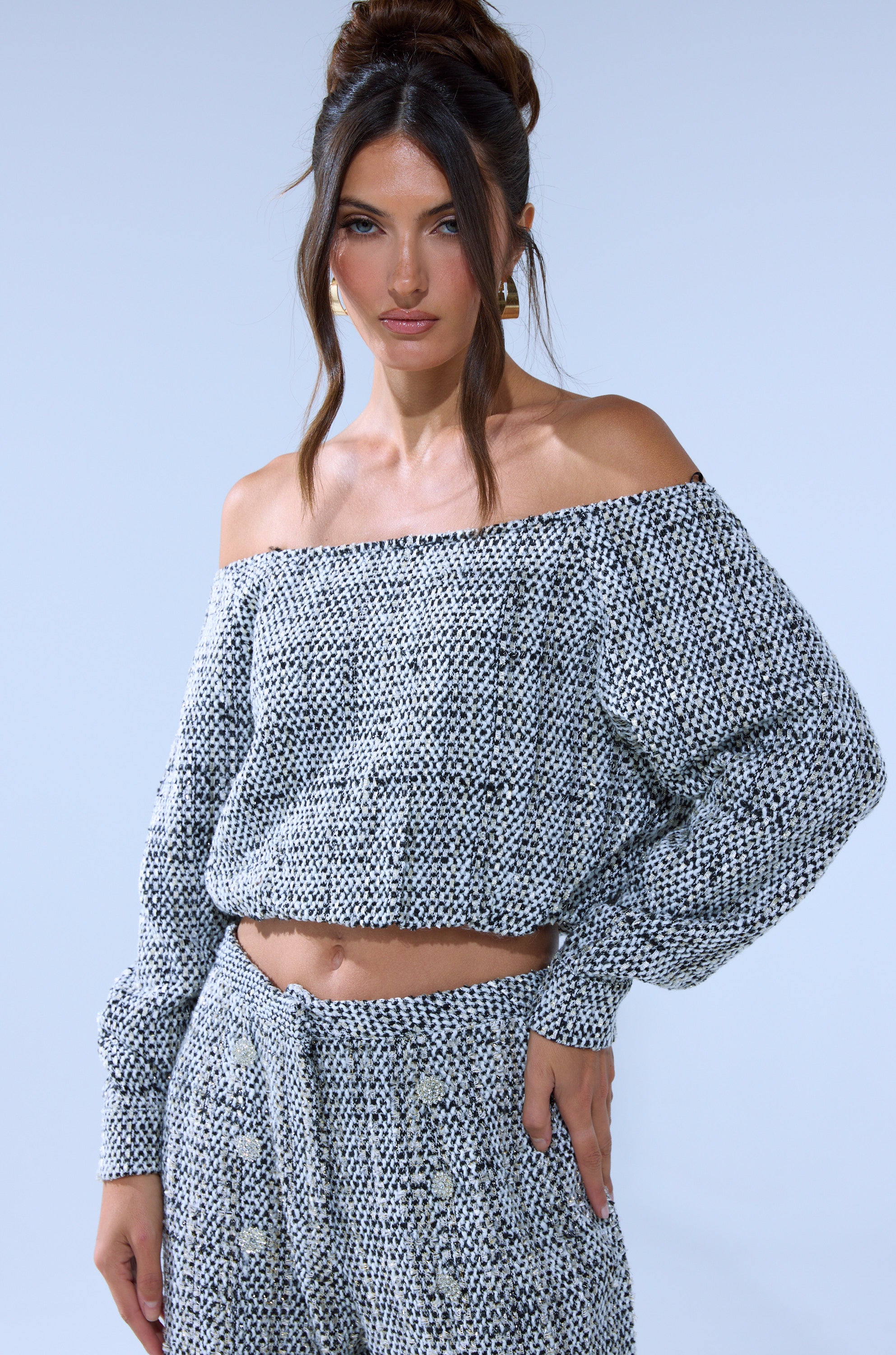 A woman with long dark hair in an updo wears the DRESSY VIBES OFF THE SHOULDER SWEATSHIRT and matching high-waisted pants, standing against a light blue background and looking confidently at the camera.