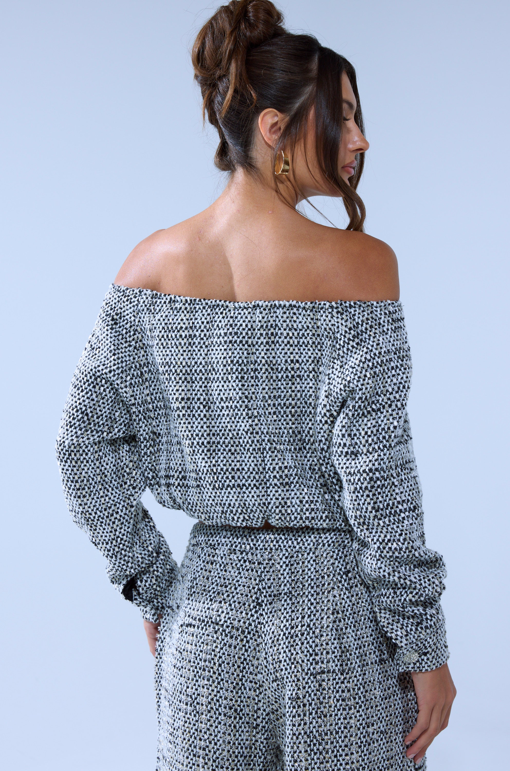 A woman with brown hair in a loose bun models the DRESSY VIBES OFF THE SHOULDER SWEATSHIRT and matching pants, posing with her back to the camera against a plain light background.