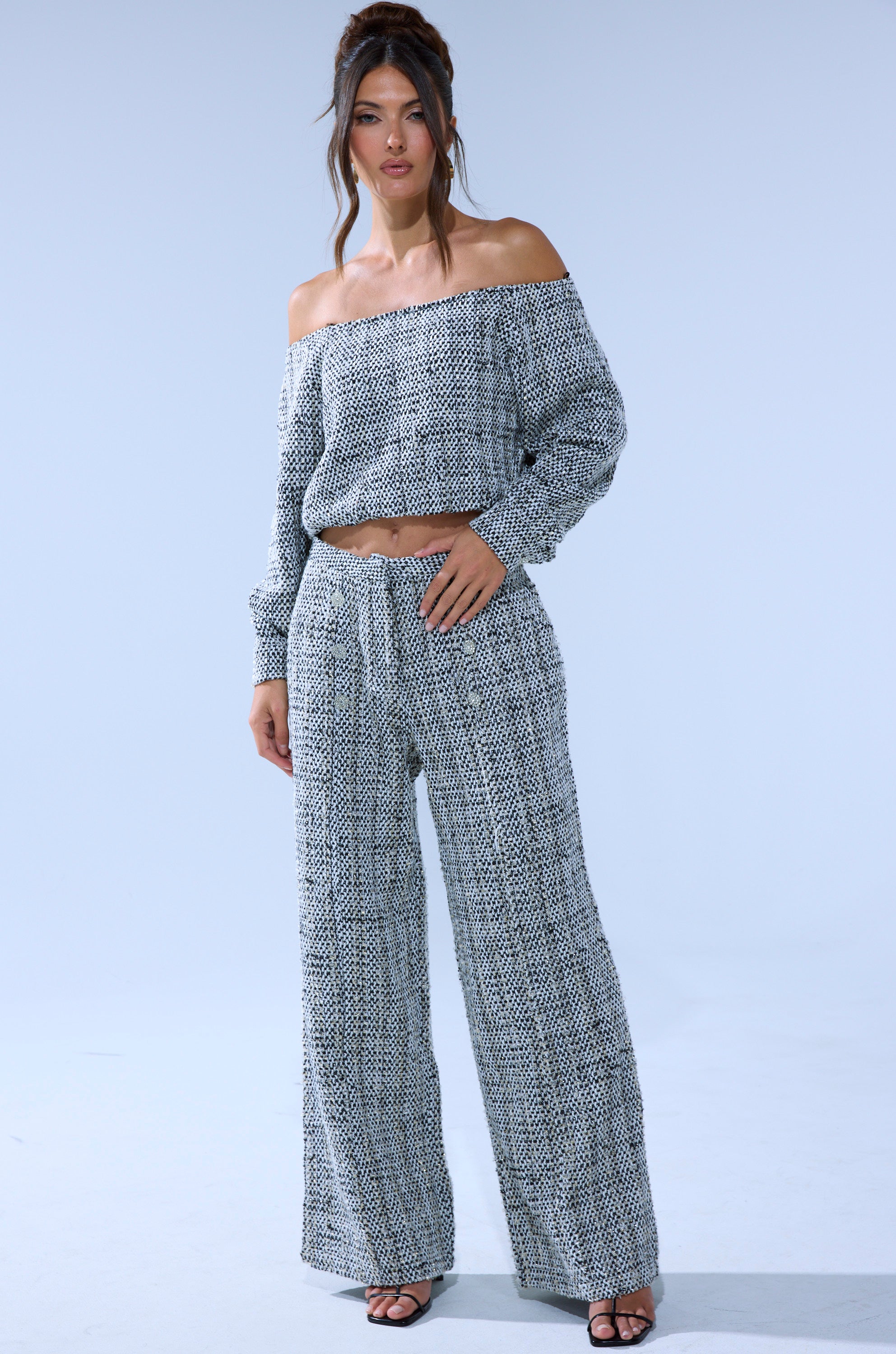A woman stands against a light blue background wearing the DRESSY VIBES OFF THE SHOULDER SWEATSHIRT and matching black and white patterned wide-leg pants, paired with black heeled sandals.