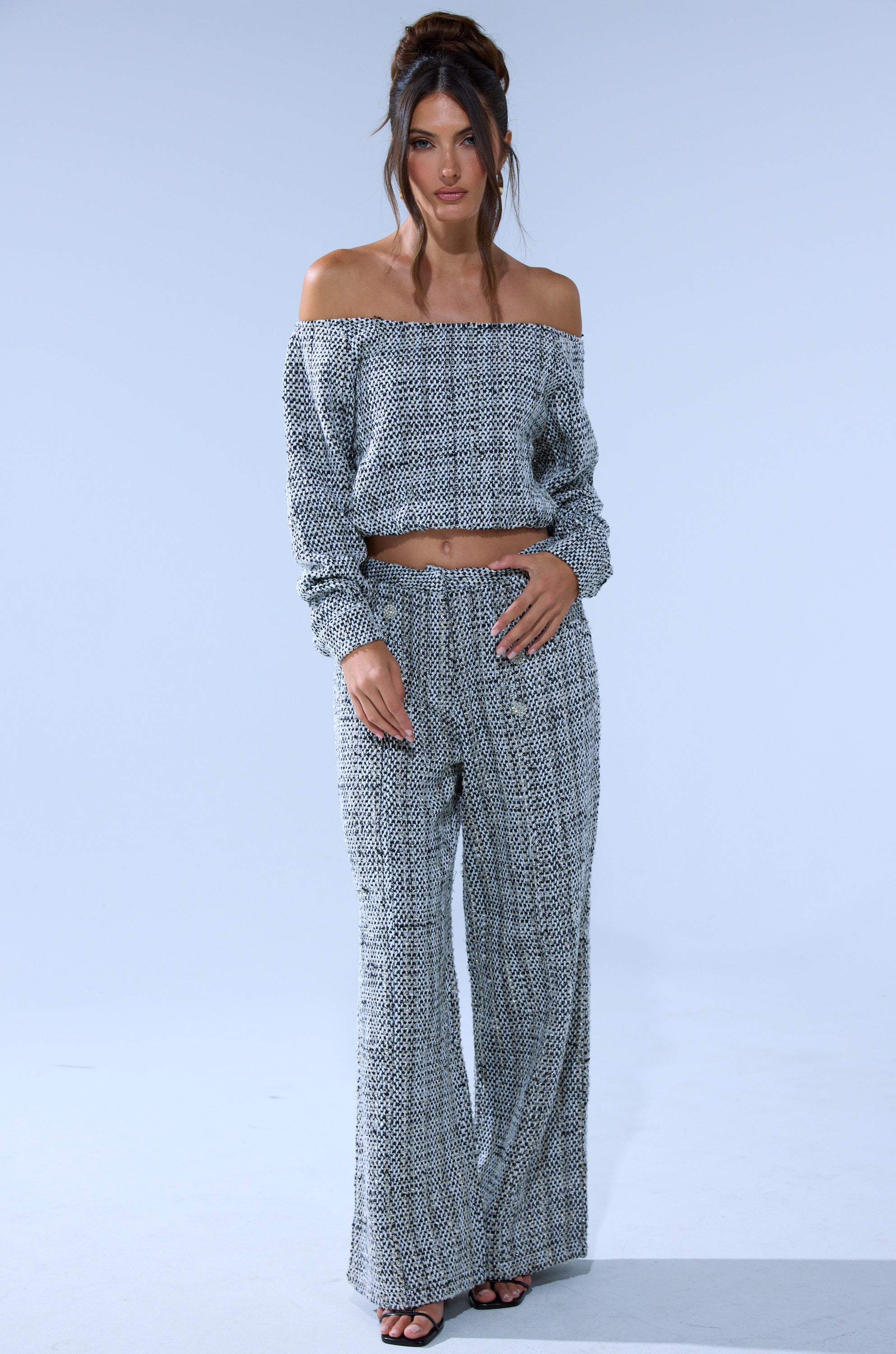 A woman poses against a light blue background in the DRESSY VIBES OFF THE SHOULDER SWEATSHIRT, paired with wide-leg pants and chic black sandals. Her long hair is styled up for a polished look.