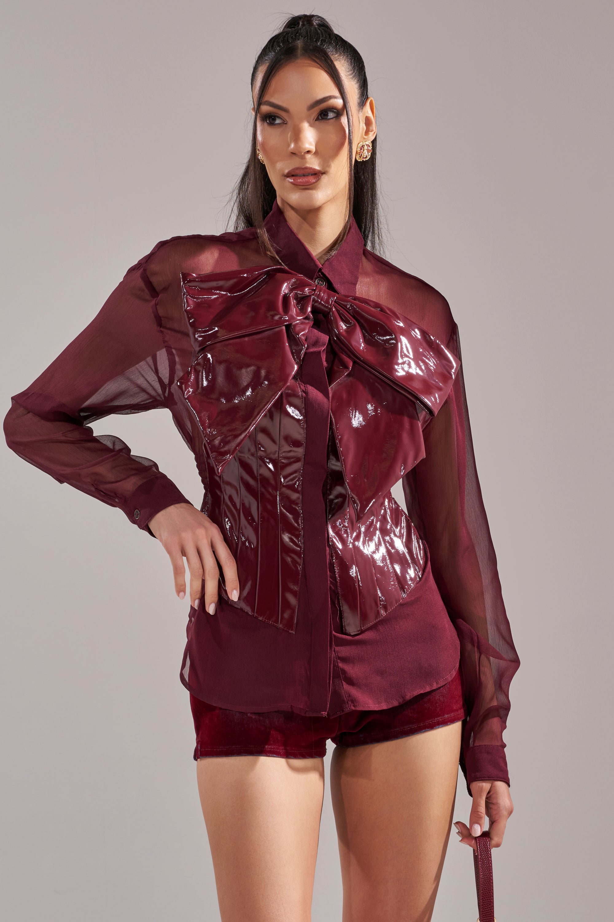 A woman models the CRAZY FOR YOU CHIFFON BUTTON DOWN featuring a sheer design, shiny oversized bow, and front detailing, paired with matching burgundy shorts against a plain light backdrop.