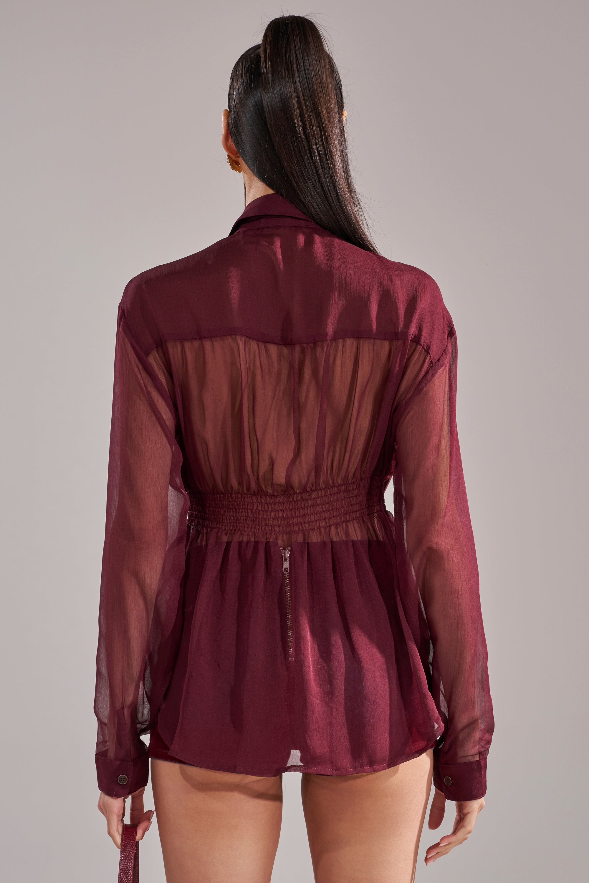 A woman with a high ponytail is shown from behind wearing the CRAZY FOR YOU CHIFFON BUTTON DOWN, a sheer burgundy blouse with a cinched waist, against a plain gray background.
