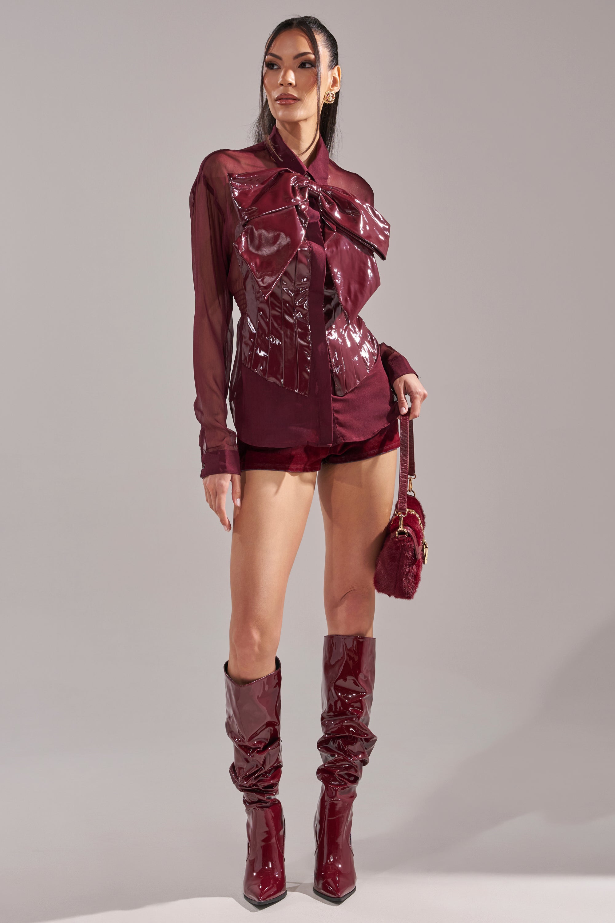 A woman poses in the CRAZY FOR YOU CHIFFON BUTTON DOWN blouse with a matching mini skirt, ruched knee-high boots, and a small fur-accented handbag, set against a plain gray background.