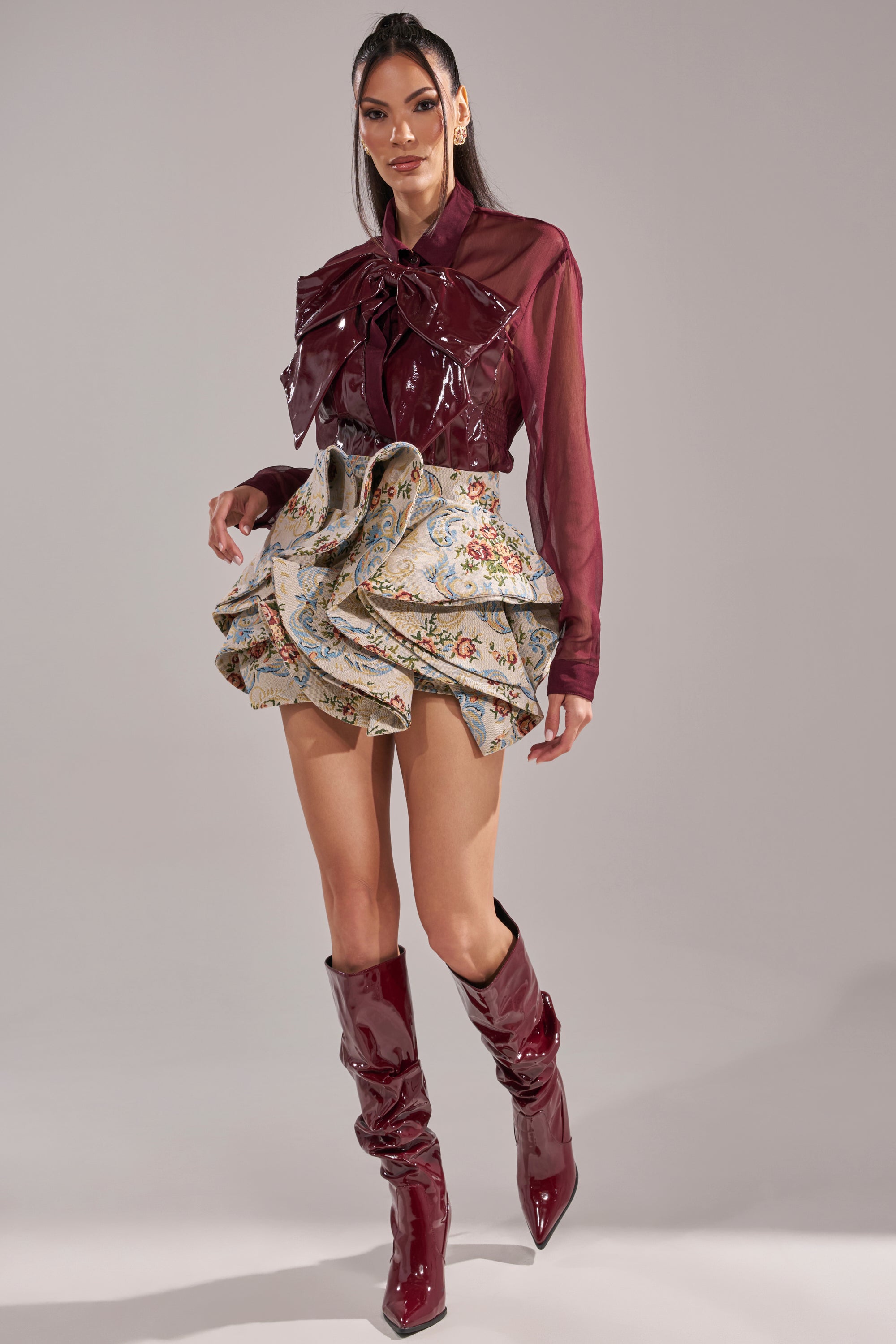 A woman poses in the CRAZY FOR YOU CHIFFON BUTTON DOWN, styled with a large bow, a ruffled floral mini skirt, and glossy burgundy knee-high boots, against a neutral background.