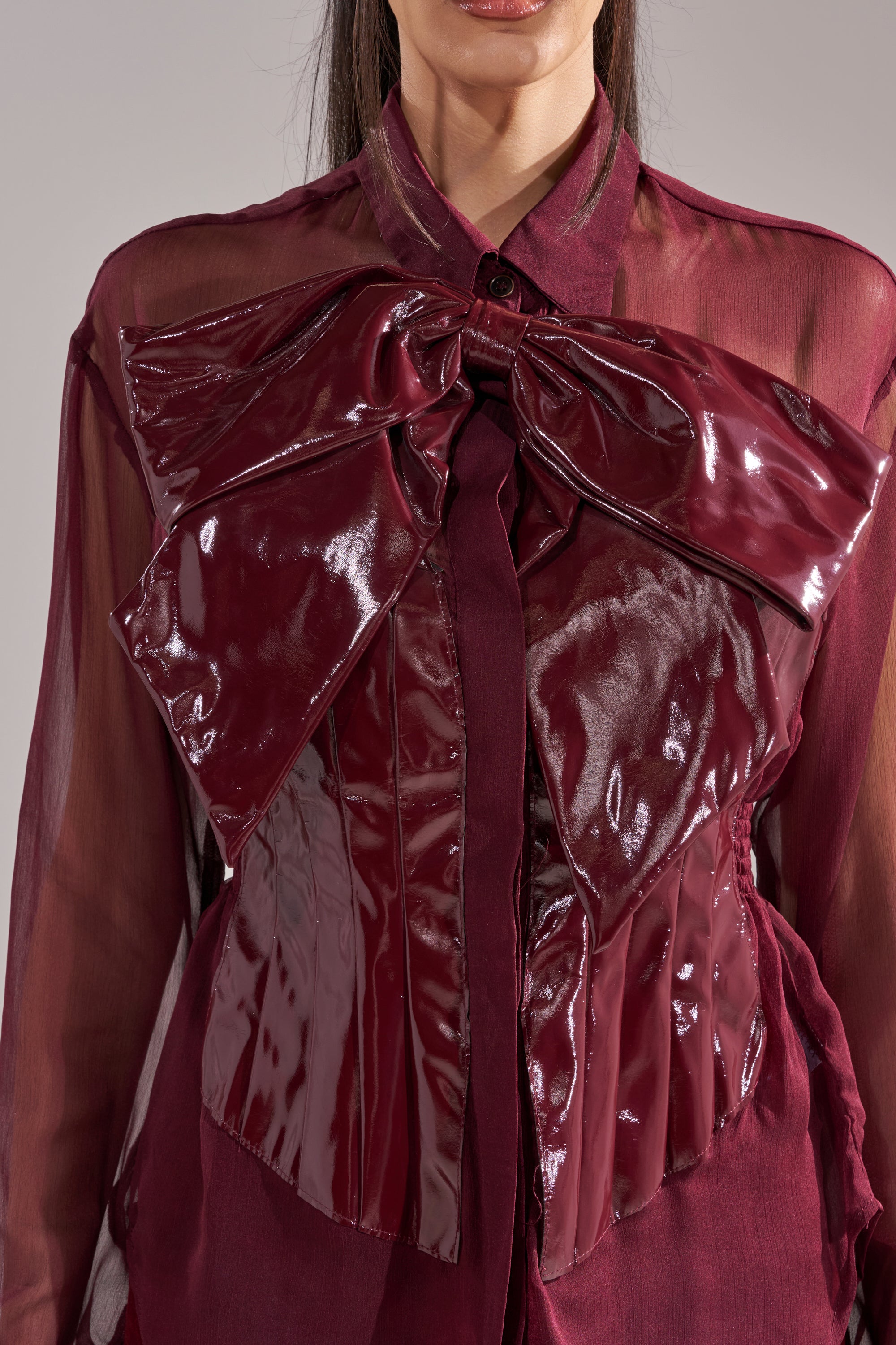 A woman models the CRAZY FOR YOU CHIFFON BUTTON DOWN, a sheer burgundy blouse with a shiny, oversized faux leather bow and corset detail on the front. The image is cropped from her shoulders to waist.