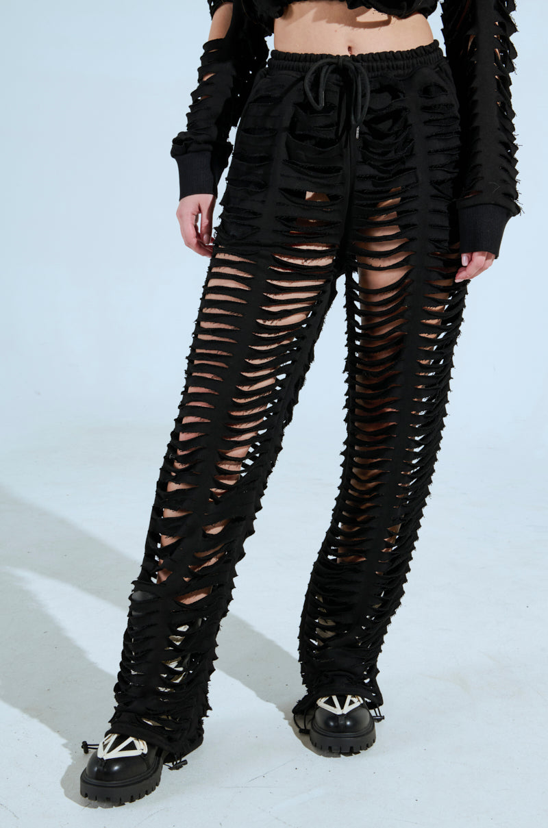 A person wears CLEAN LINES SLASHED PANT with an AKIRA Label top, both featuring numerous slits. The ensemble is complemented by black shoes with metallic accents, set against a plain white background. 1