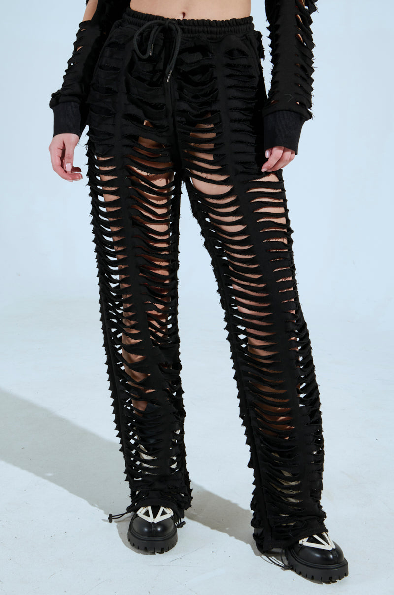 Sporting the edgy AKIRA Label style, a person wears black CLEAN LINES SLASHED PANT featuring horizontal cuts and a matching top. The high-rise pants paired with chunky black shoes create a bold look against a plain backdrop. 3