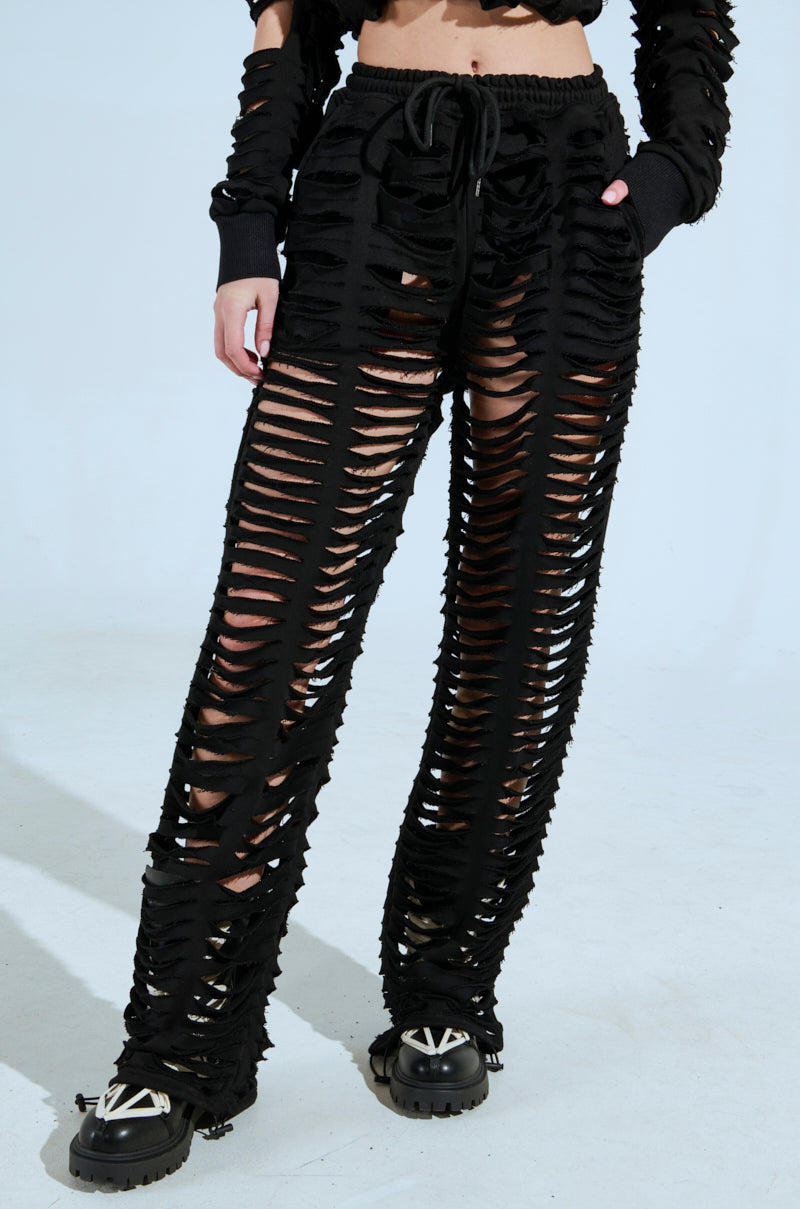 A person sporting the edgy style of the CLEAN LINES SLASHED PANT in black with heavy distressing reveals skin through horizontal slashes. They complement the look with a matching long-sleeve top and black platform shoes adorned with decorative elements. 4