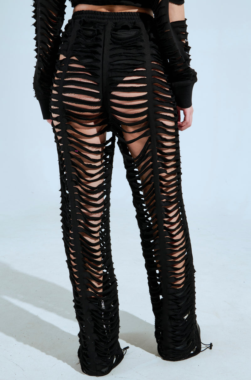 A person models the CLEAN LINES SLASHED PANT by AKIRA Label, featuring trendy black fabric with horizontal slits exposing skin and a stylish high-rise fit, against a plain background. 6