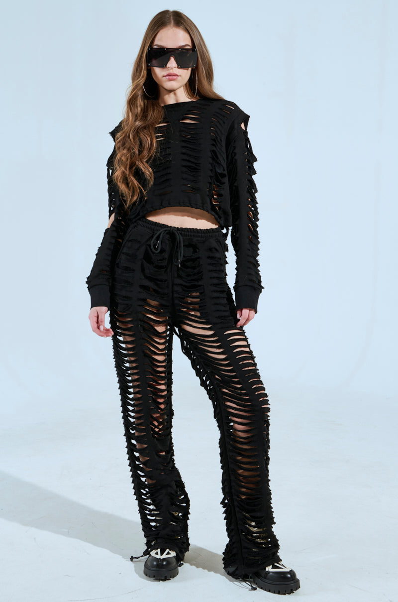 A person confidently poses against a plain backdrop, wearing large sunglasses and a black distressed two-piece outfit with the CLEAN LINES SLASHED PANT from the AKIRA Label. Their long, wavy hair and black platform shoes complete the ensemble. 7