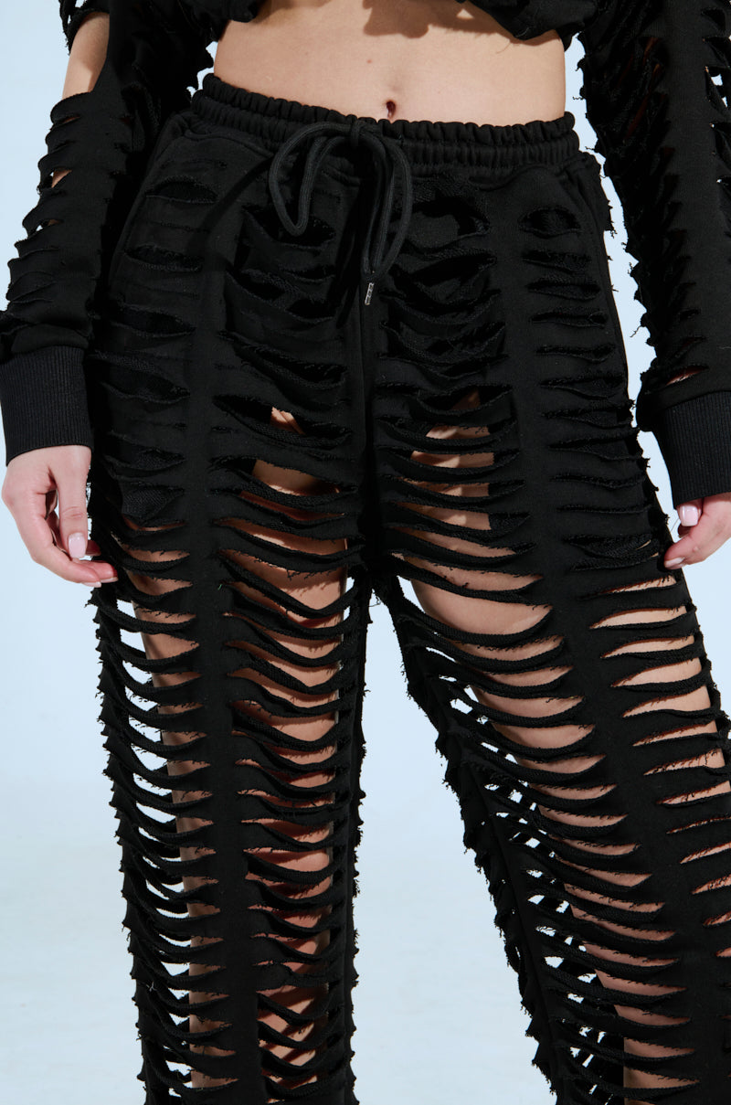 A person wearing AKIRA Label's CLEAN LINES SLASHED PANT, featuring a high-rise black design with slashed fabric from waist to ankles, reveals skin through the gaps. This striking look is perfectly paired with a matching top for an edgy, bold statement. 8