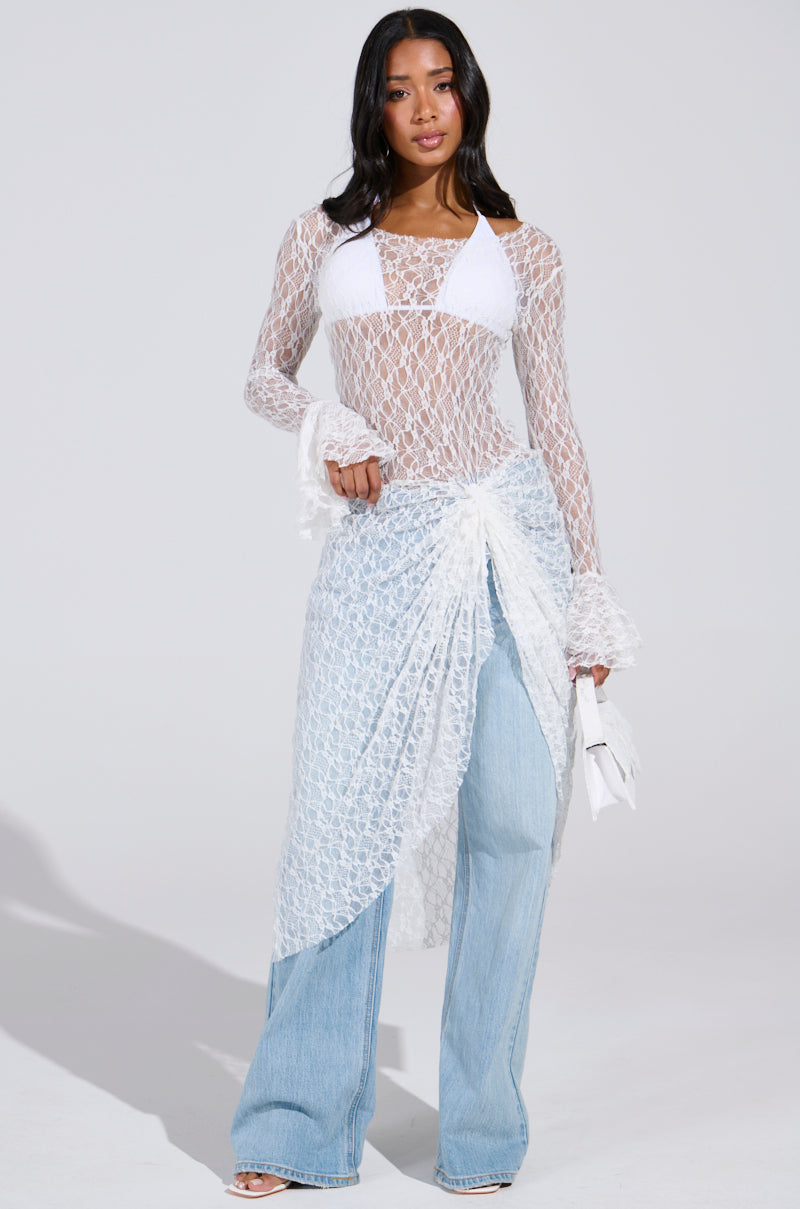 A woman poses festival-ready in a LILY LACE BLOUSE IN WHITE over a bikini top, paired with light blue jeans and a sheer lace skirt. She holds a small white bag, exuding boho-chic vibes against a plain light gray backdrop.
