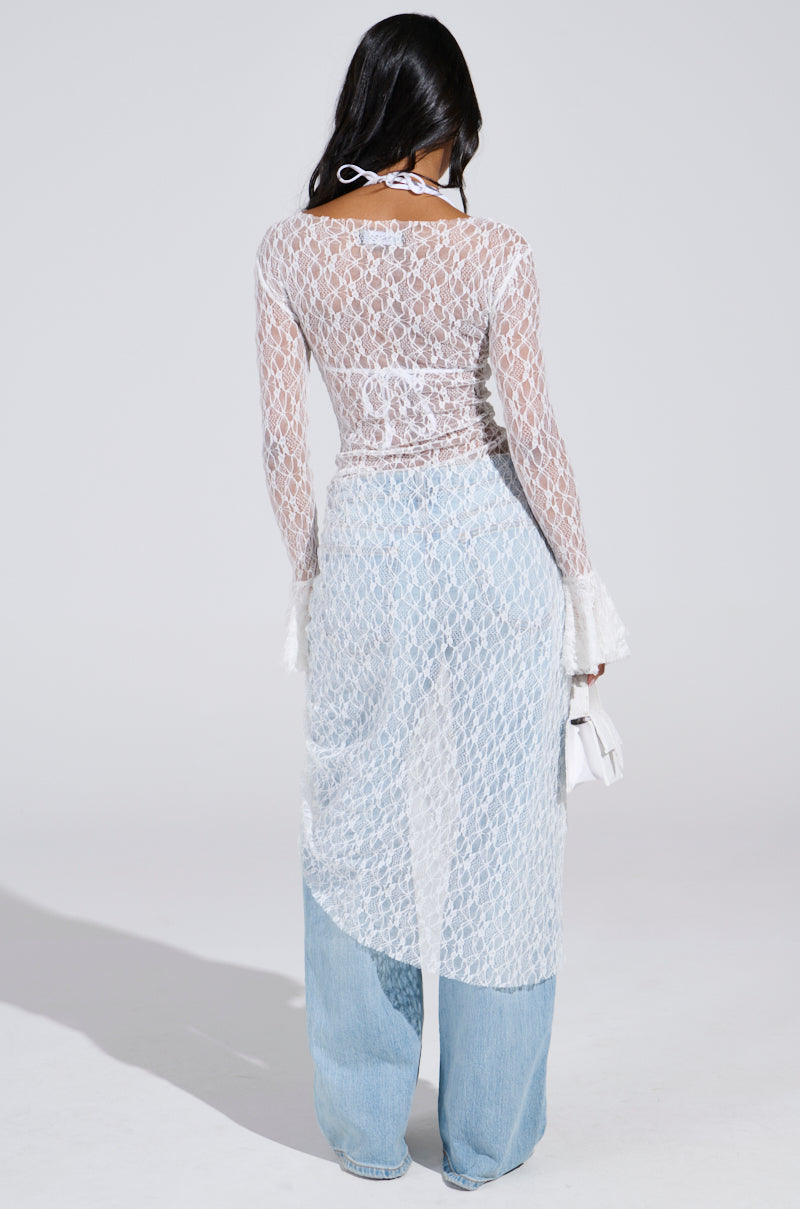 Person with long dark hair wears a LILY LACE BLOUSE IN WHITE over light blue jeans, exuding boho chic vibes. Holding a small white handbag, their head is turned slightly to the side.