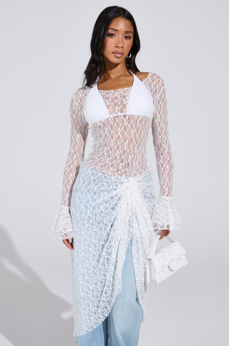 A woman exudes boho chic style in a sheer Lily Lace Blouse in White over a bikini, paired with light blue jeans and a delicate lace wrap skirt. Holding a small lace purse, she poses against a plain background. Festival-ready, her look is effortlessly elegant.