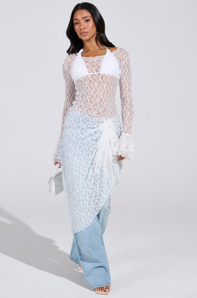 A woman confidently strolls in a LILY LACE BLOUSE IN WHITE with a sheer skirt overlay, exuding boho chic. She pairs it with light blue jeans and white heels, carrying a small white bag against a plain light gray backdrop.