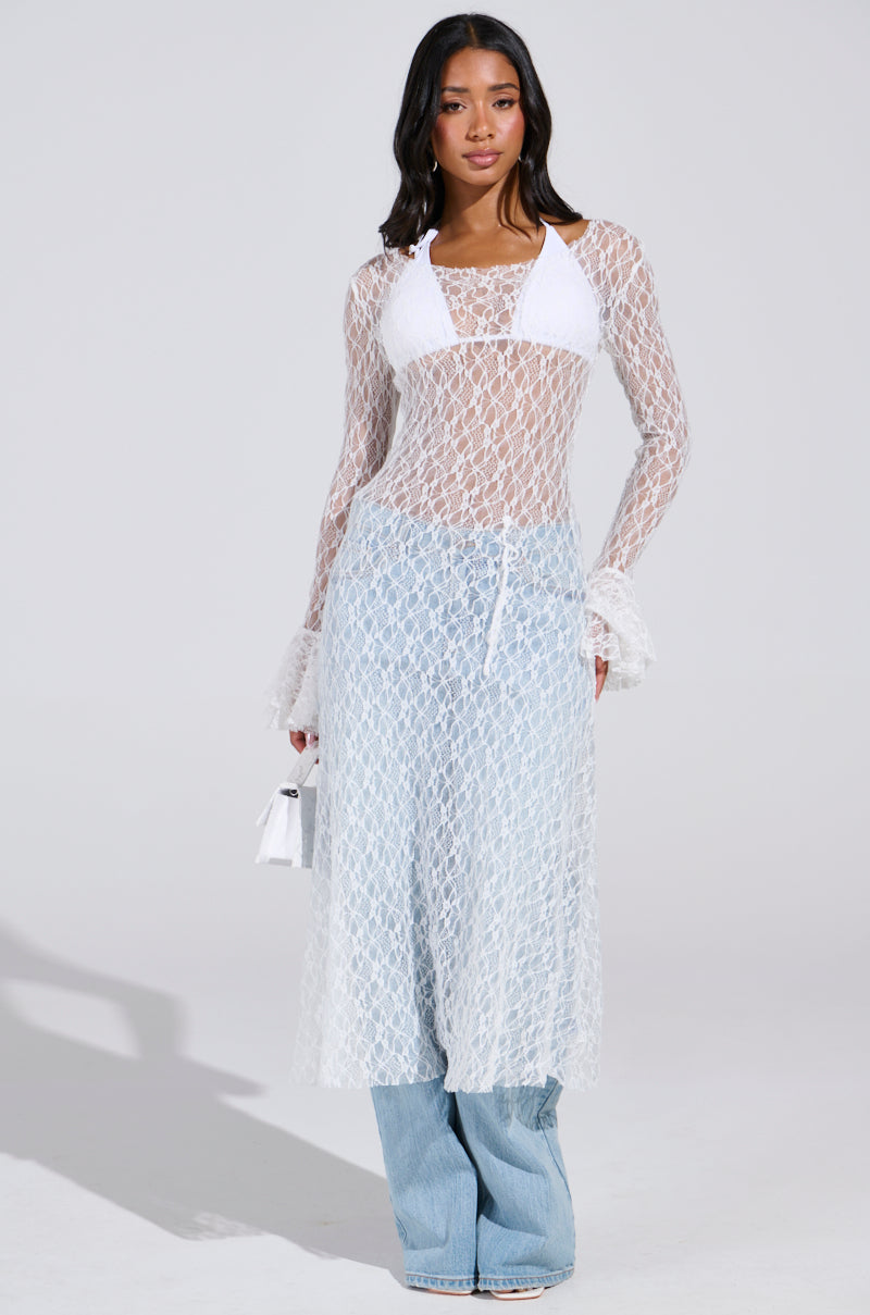 A woman wears a LILY LACE BLOUSE IN WHITE over a bikini top and light blue jeans, holding a small white purse with boho chic style. Her dark hair is loosely styled against the plain white background.