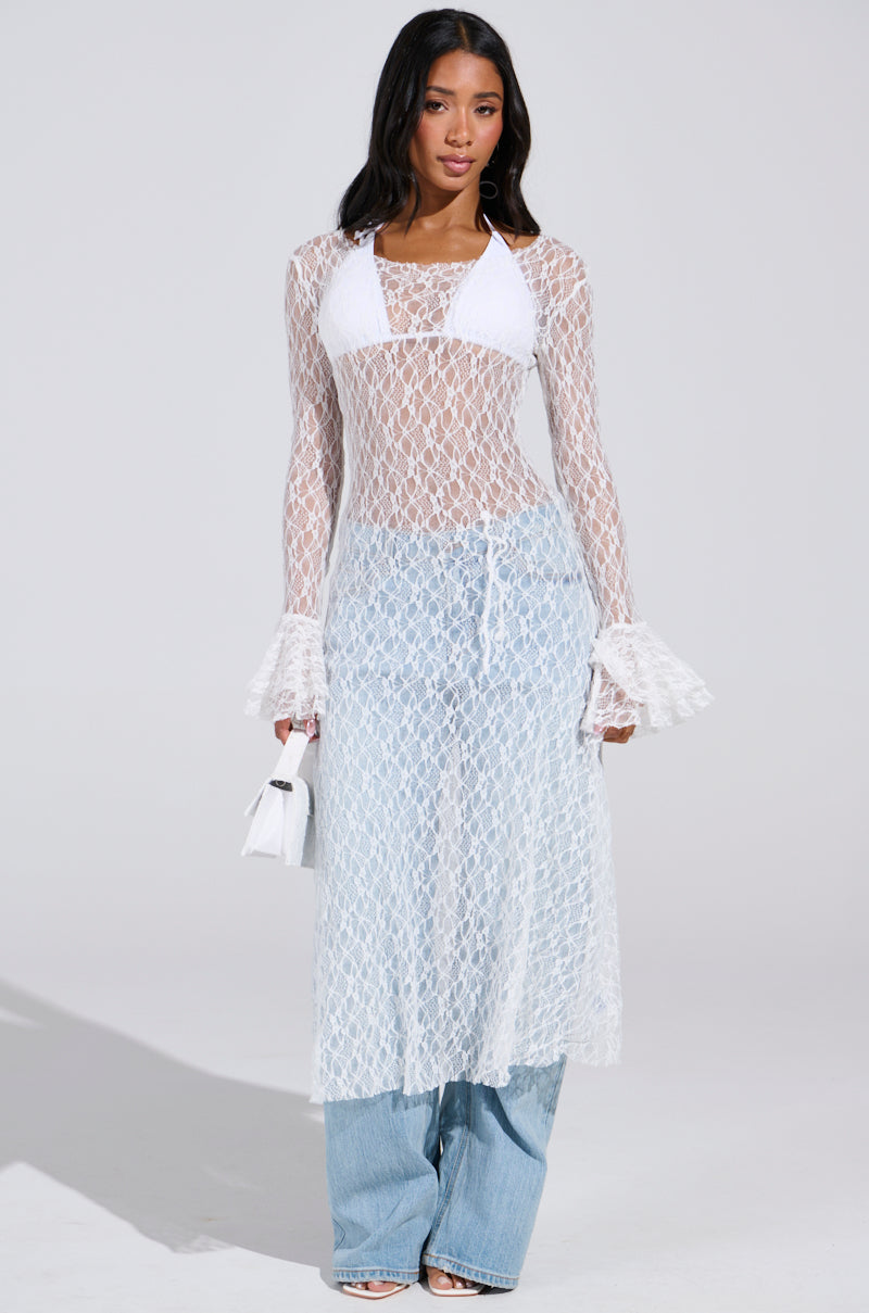 In a well-lit studio, a woman exudes boho chic style in a sheer LILY LACE BLOUSE IN WHITE over a bikini top and blue jeans. She holds a small white purse, with the blouse's long sleeves and flared cuffs enhancing her effortlessly stylish look.