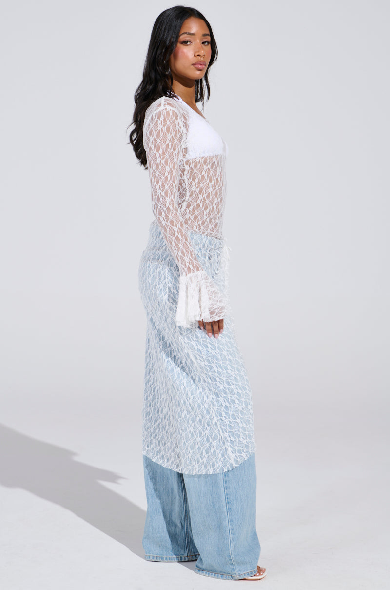A woman poses in a stylish ensemble featuring the LILY LACE BLOUSE IN WHITE and light blue wide-leg jeans. She stands in profile against a plain background, her long dark hair cascading past her shoulders, exuding boho chic elegance.