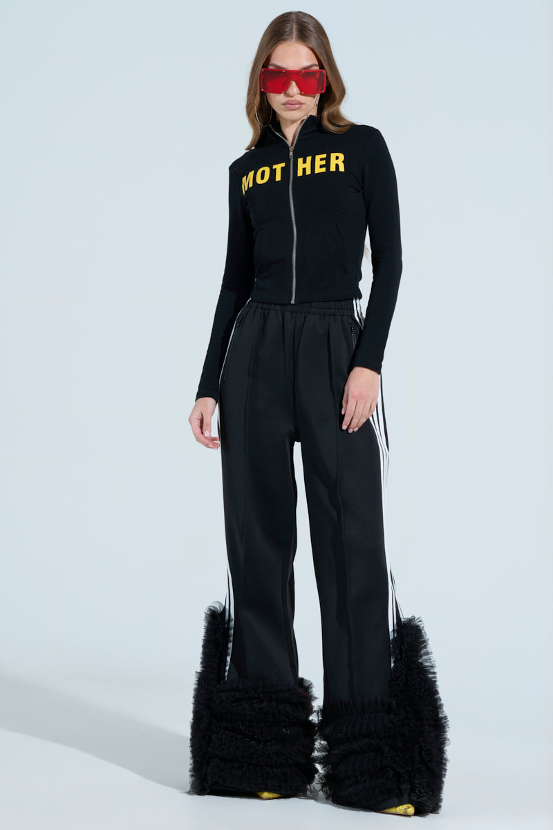 A woman in black, wearing an AKIRA Label MOTHER F*UCKER zip-up sweatshirt with "MOTHER" bold in yellow. She pairs it with black pants featuring white stripes and fluffy leg coverings. Her red sunglasses add flair as she poses confidently against a light background.