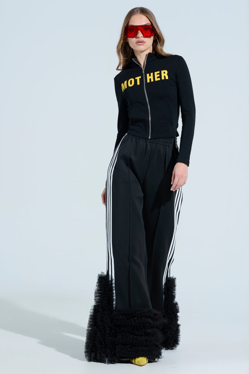 Against a light blue backdrop, a woman wears red sunglasses and the MOTHER F*UCKER ZIP UP SWEATSHIRT, paired with black pants featuring white stripes and fluffy hems. She channels the AKIRA Label vibe as she poses confidently with one hand on her hip.