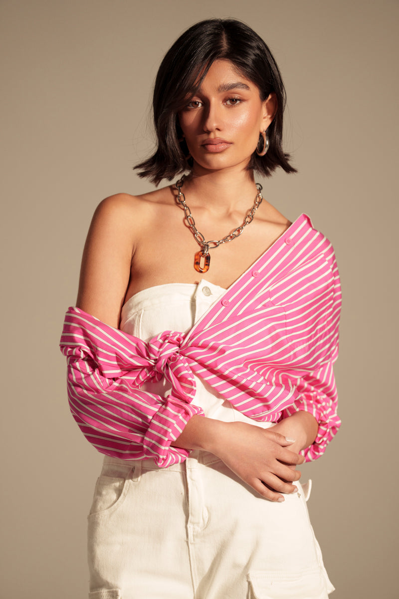 A woman with short dark hair wears the CARLY BUTTON DOWN, a dual-tone striped shirt styled off one shoulder and tied at the waist over a white strapless top and light pants, accessorized with a chunky chain necklace.