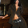 A woman with long dark hair exudes confidence in chic fall fashion, wearing the FALL VIBES VELVET DENIM BUTTON DOWN and flared pants by a wooden bar, highlighting her decorative belt and stylish look.