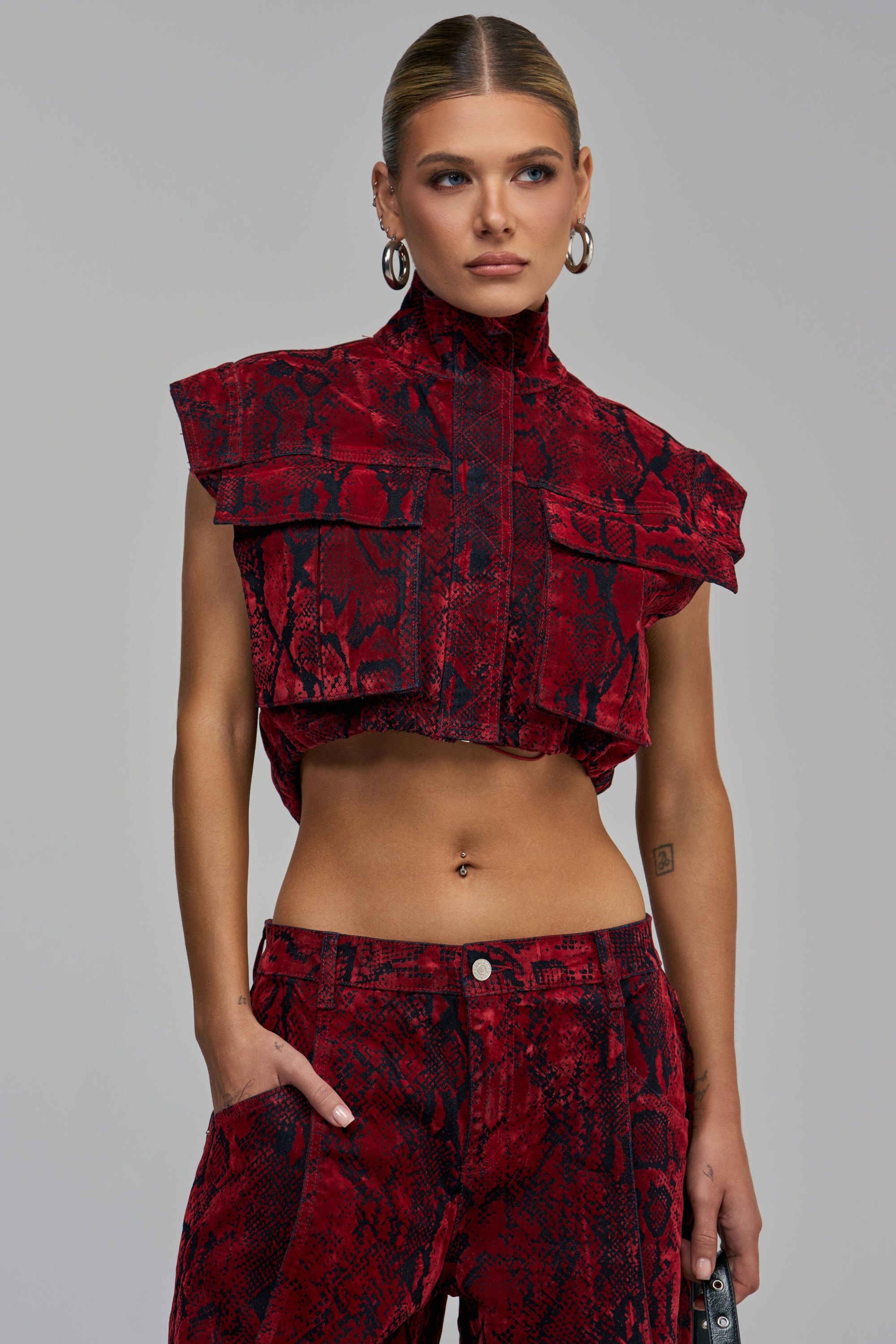 A person wearing the TEXTURED TEMPTATION BUTTON DOWN, a sleeveless, cropped red and black snakeskin velvet top with large chest pockets and matching trousers.