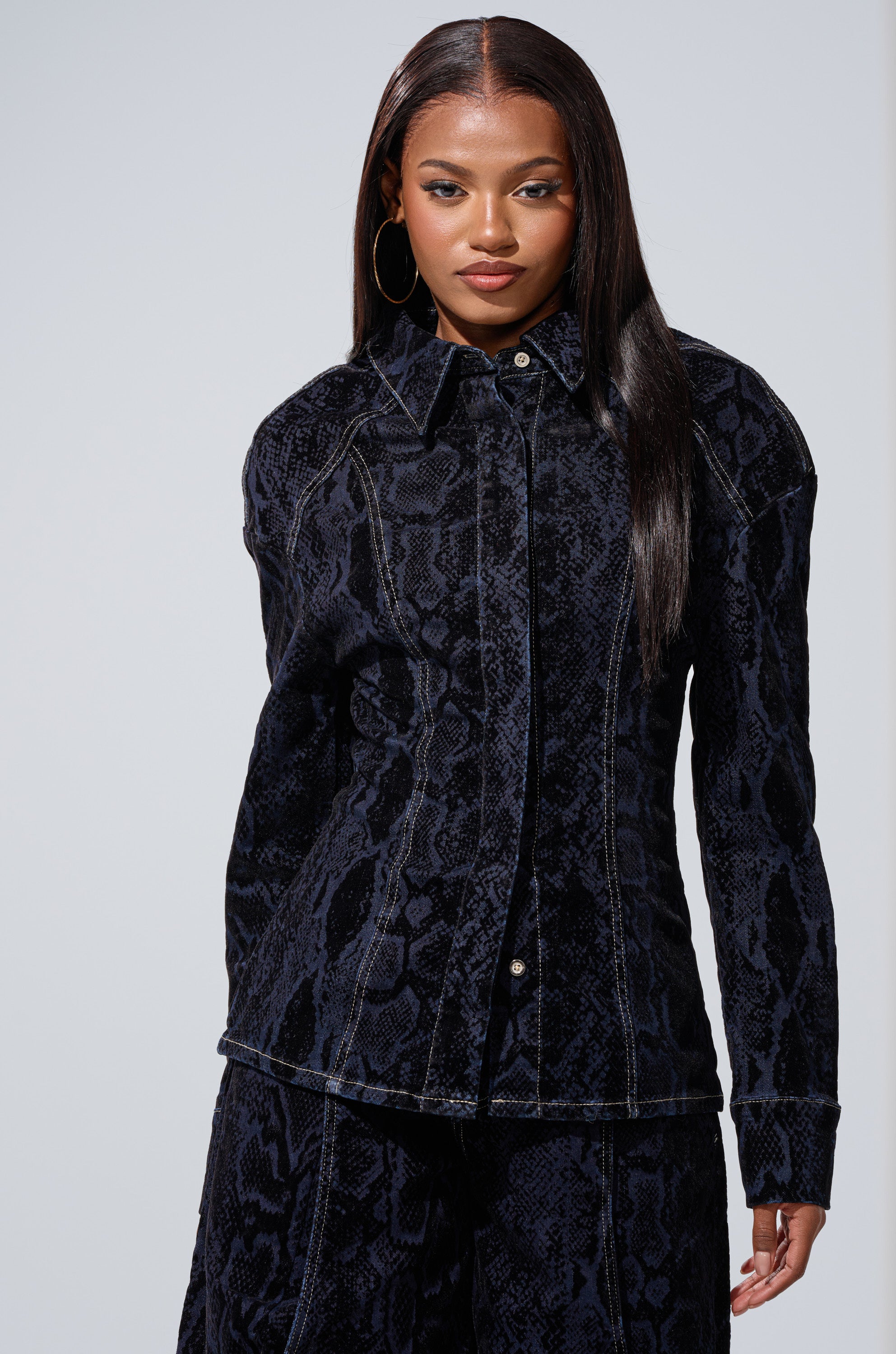 A woman with long straight hair wears the FEELS LIKE MONEY DENIM BUTTON DOWN and matching pants. She stands facing forward, hands at her sides, showcasing hoop earrings and a confident expression.