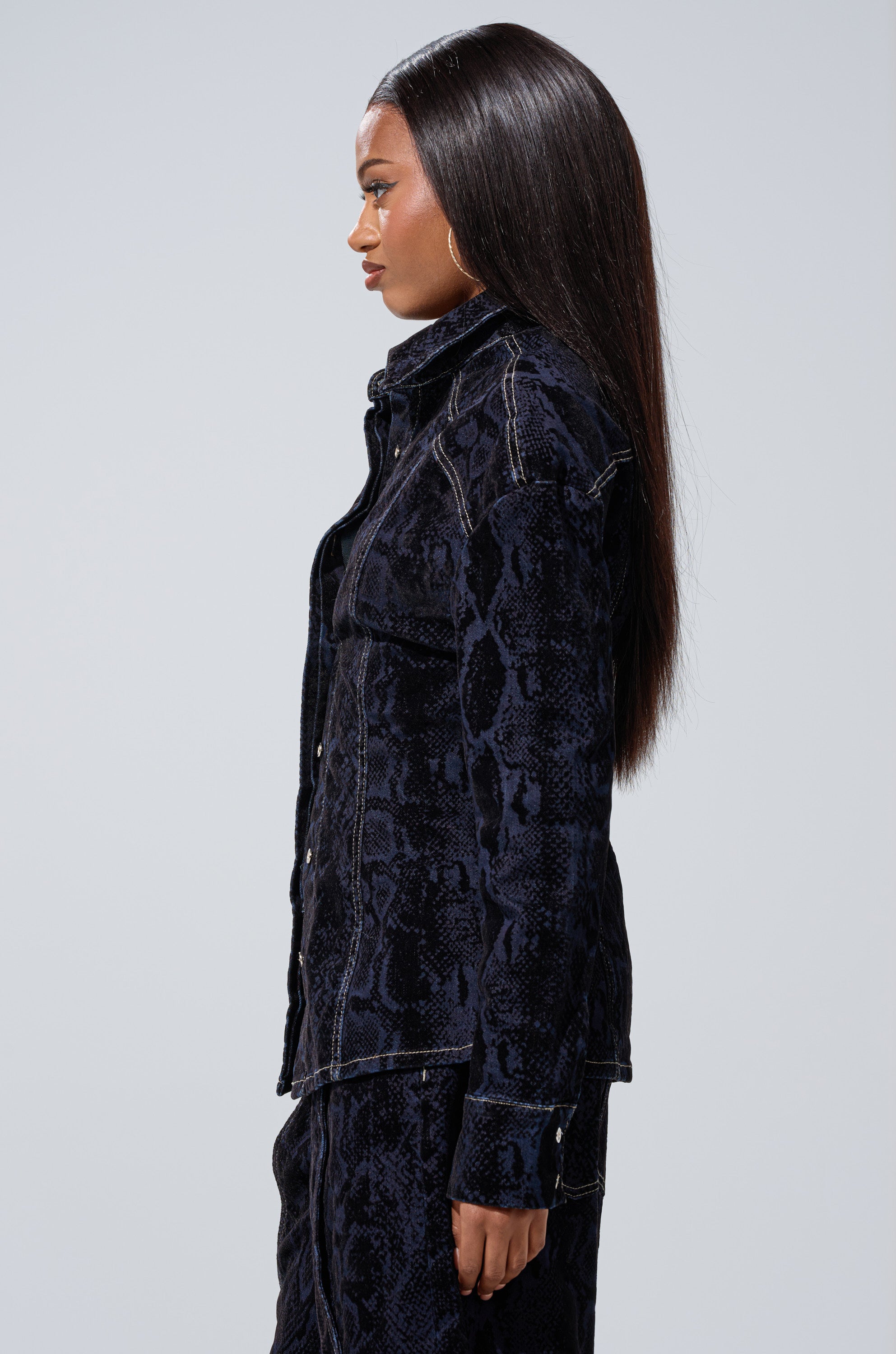 A woman with long, straight hair stands in profile against a plain light background, wearing the FEELS LIKE MONEY DENIM BUTTON DOWN and matching curve-hugging dark-wash jeans.