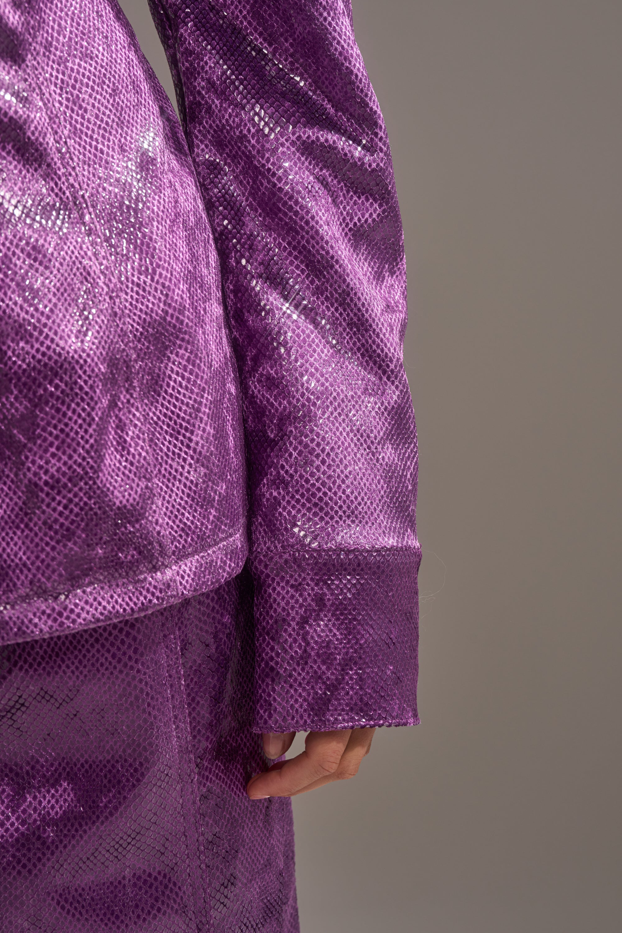 Close-up of a person wearing the AKIRA Label CRAZY TOUCH VELVET BUTTON DOWN, a shiny purple snakeskin print top with long sleeves, shown with matching skirt against a gray background. Only the arm and part of the outfit are visible. 9