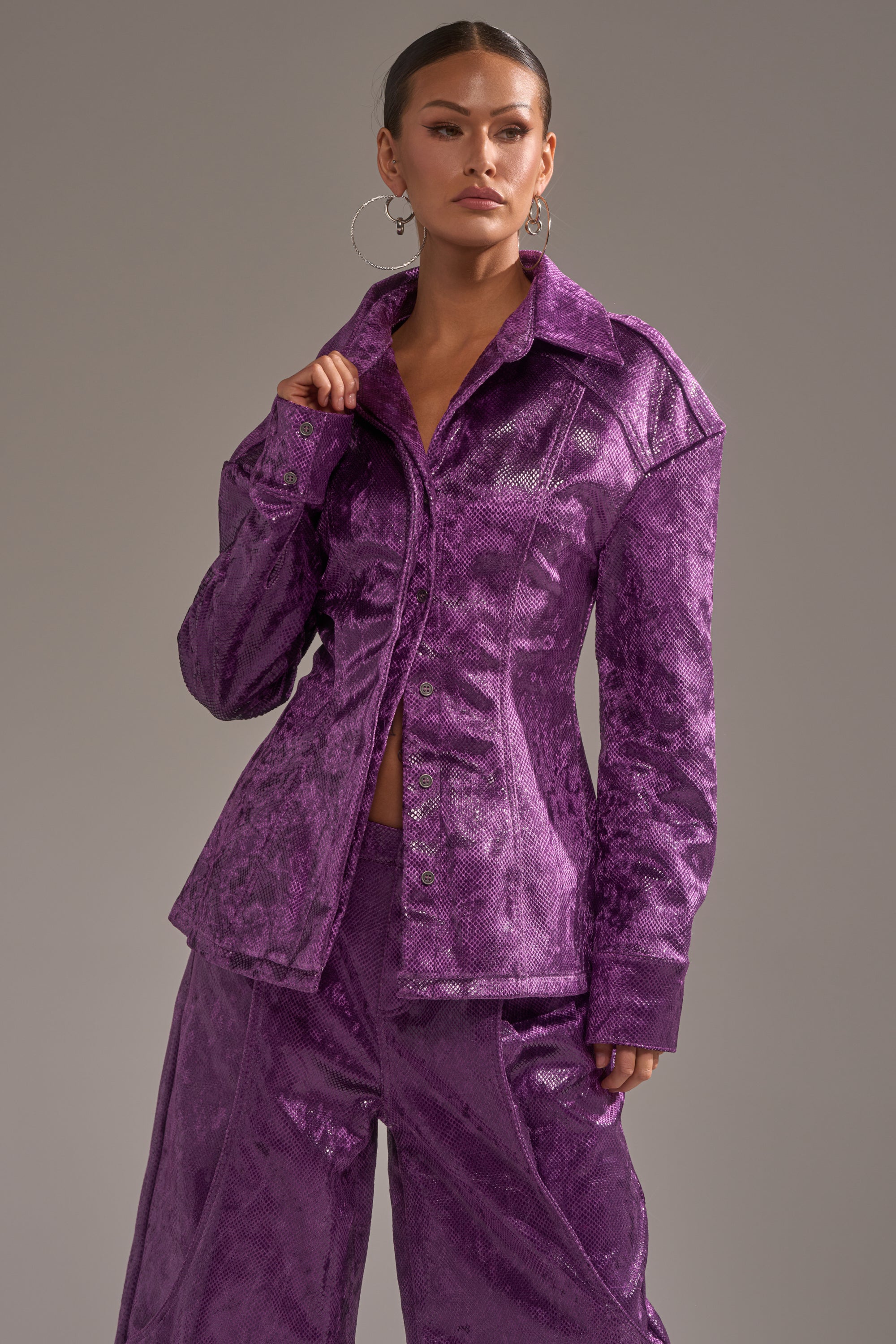 A woman poses confidently against a gray background in the shiny, textured purple CRAZY TOUCH VELVET BUTTON DOWN with matching wide-leg pants. She has slicked-back hair, large hoop earrings, and holds the jacket collar with one hand. 3