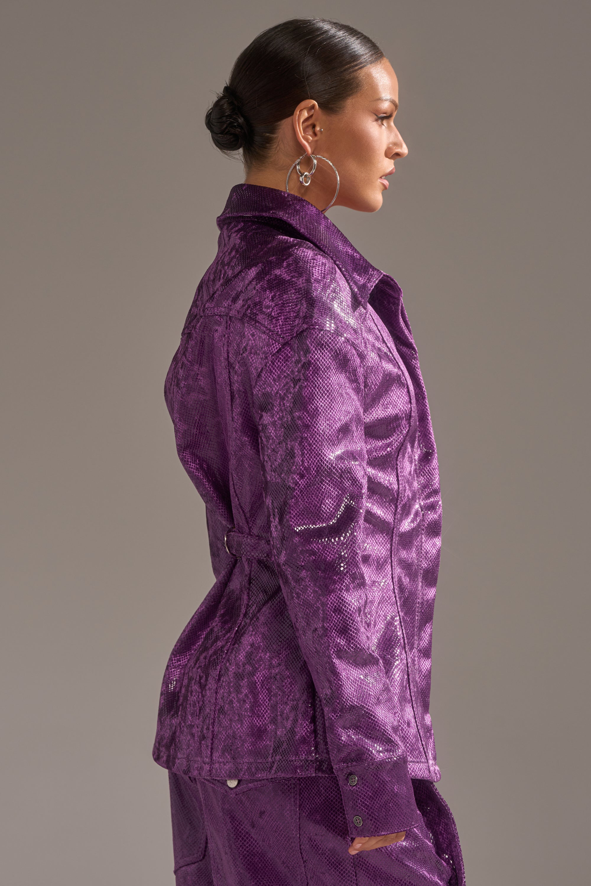 A woman with slicked-back hair in a low bun wears silver hoop earrings and the shiny, textured purple CRAZY TOUCH VELVET BUTTON DOWN jacket with matching pants, standing in profile against a plain, neutral background. 4