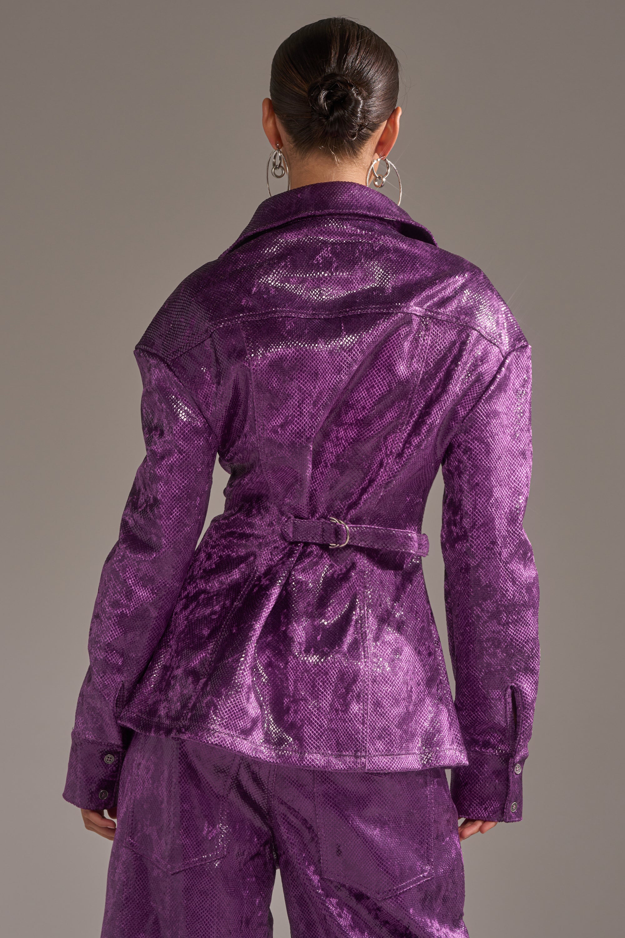 A person with dark hair in a bun is seen from the back wearing the CRAZY TOUCH VELVET BUTTON DOWN in shiny purple with a fitted longline silhouette, wide collar, belted waist, and matching pants for a modern look. 5