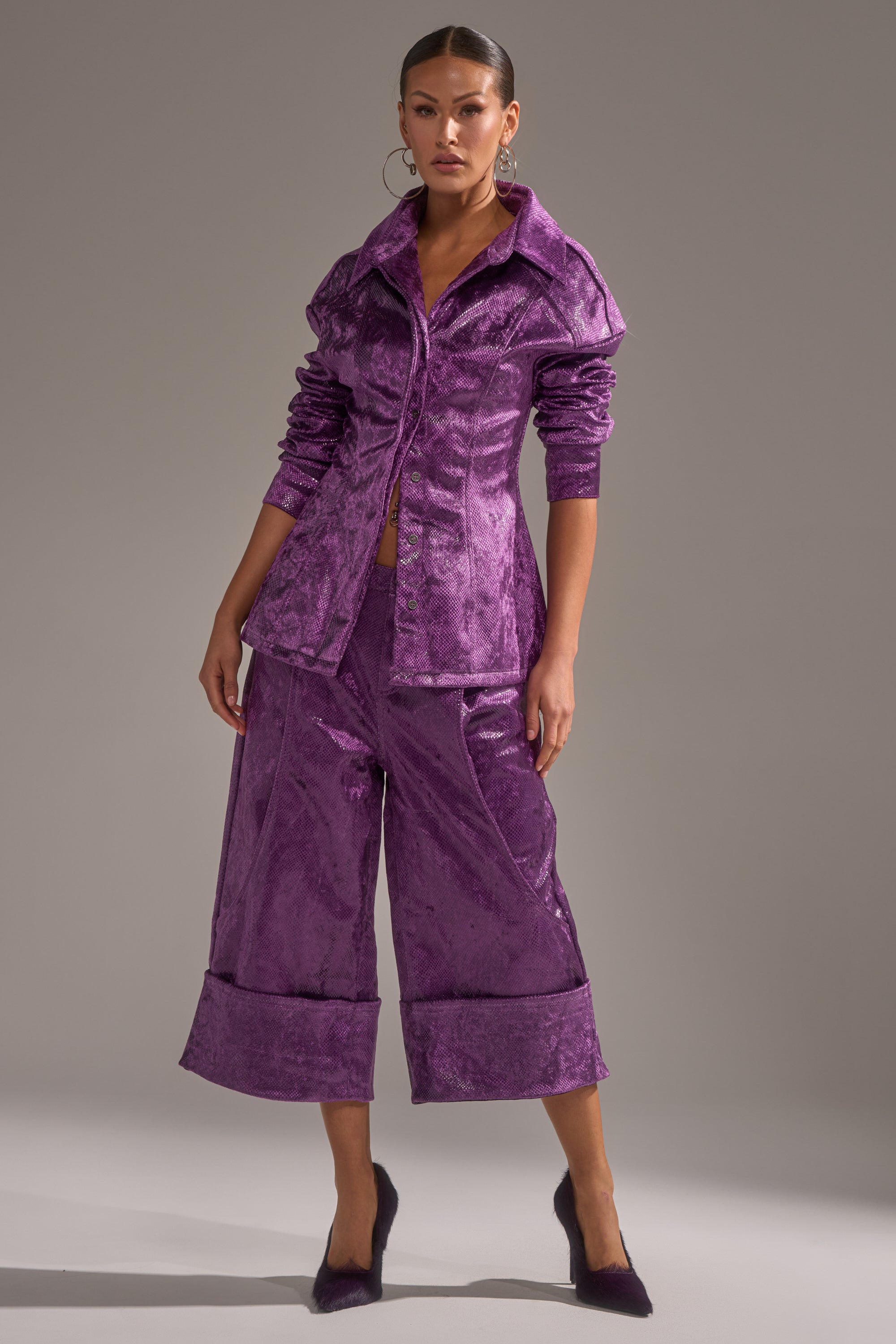 A woman stands confidently in the CRAZY TOUCH VELVET BUTTON DOWN—shiny purple, paired with wide-leg cropped pants, pointed-toe purple heels, and an AKIRA Label snakeskin print top—all set against a plain gray background. 7