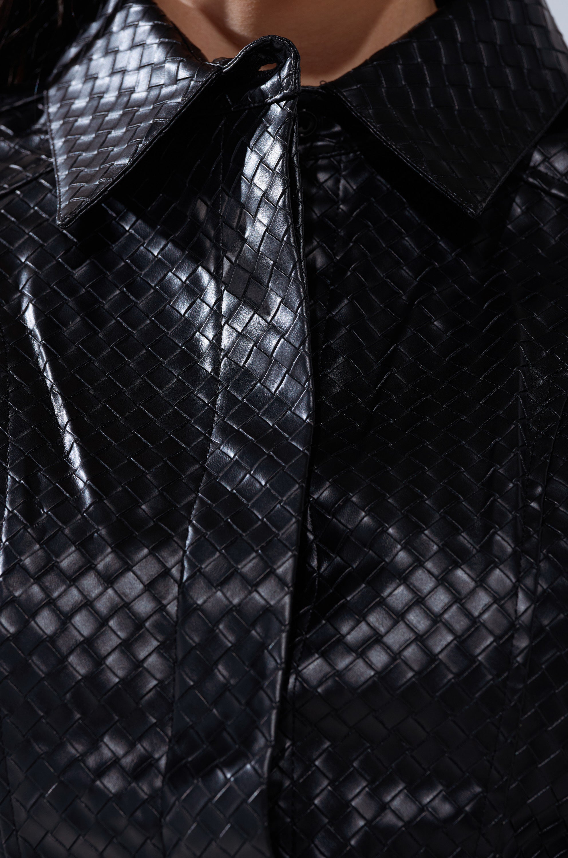 Close-up of the FEELING RICH FAUX LEATHER BUTTON DOWN IN BLACK, featuring a shiny basket-weave texture and pointed collar. The faux leather material reflects light, emphasizing its intricate design against a neutral background.