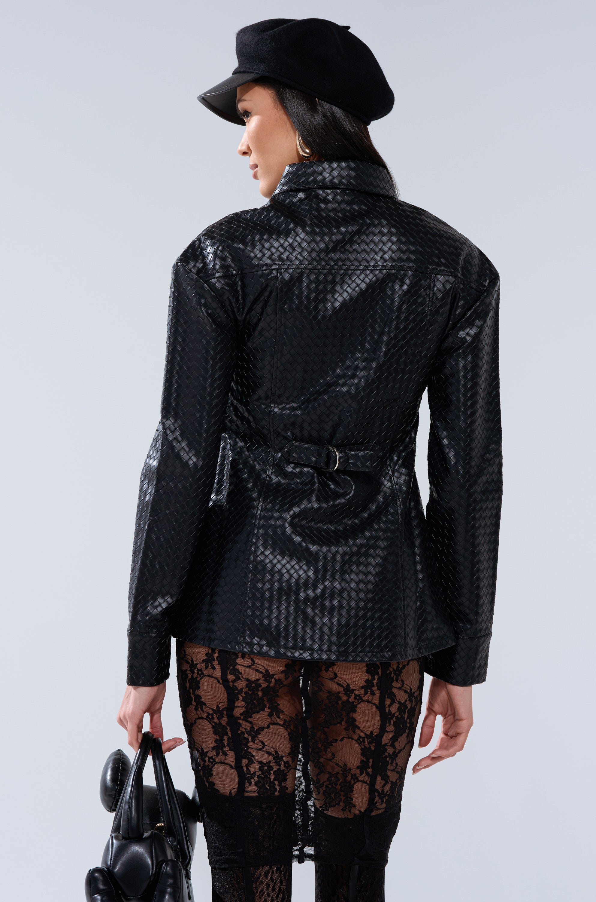 A woman is seen from behind wearing the FEELING RICH FAUX LEATHER BUTTON DOWN IN BLACK, styled as a textured jacket with a belt, black lace pants, a black newsboy cap, and holding a black handbag against a plain light gray background.