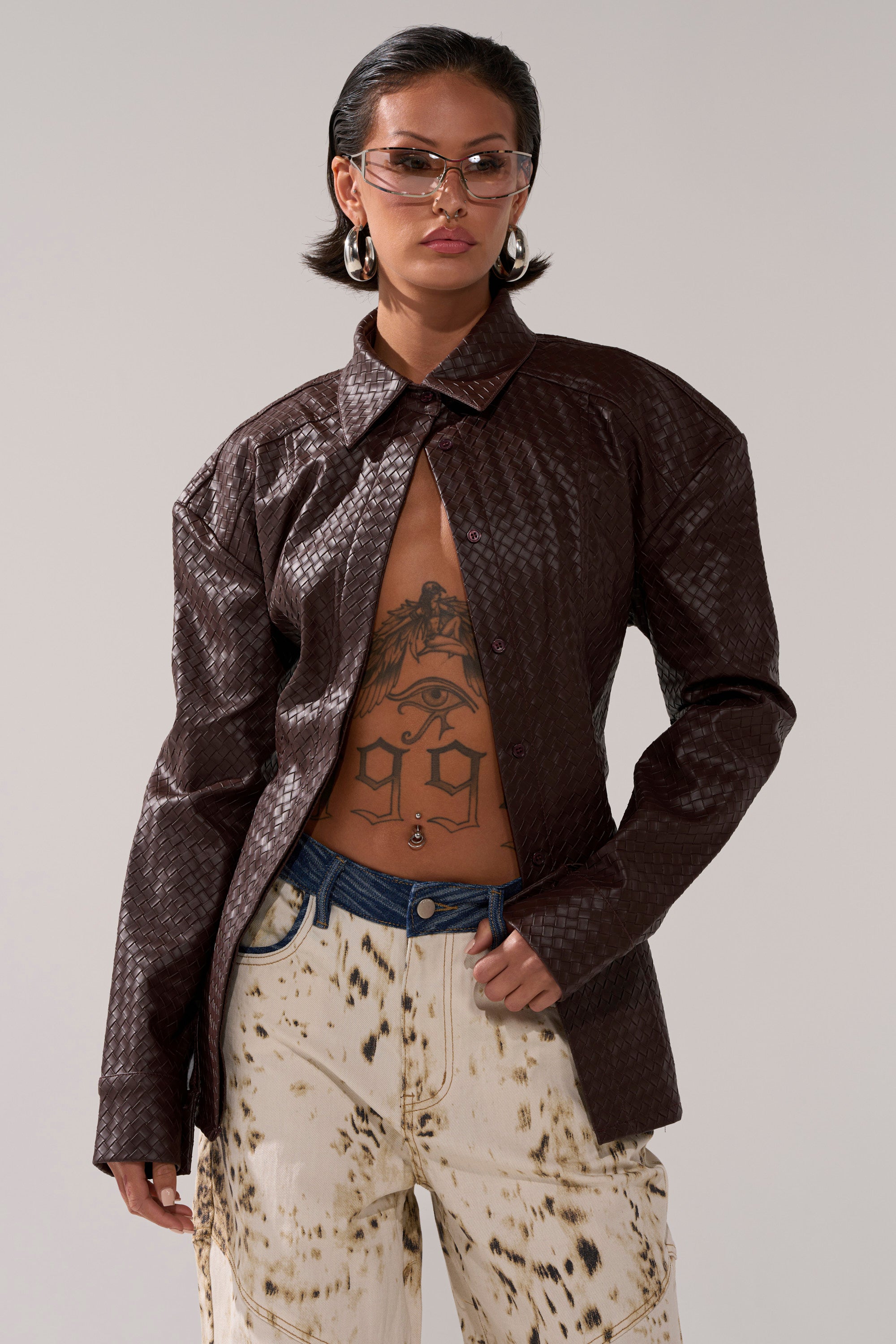 A person with short hair and glasses wears the FEELING RICH FAUX LEATHER BUTTON DOWN partly unbuttoned, revealing a torso tattoo, styled with a woven top underneath, paint-splattered jeans, and hoop earrings.